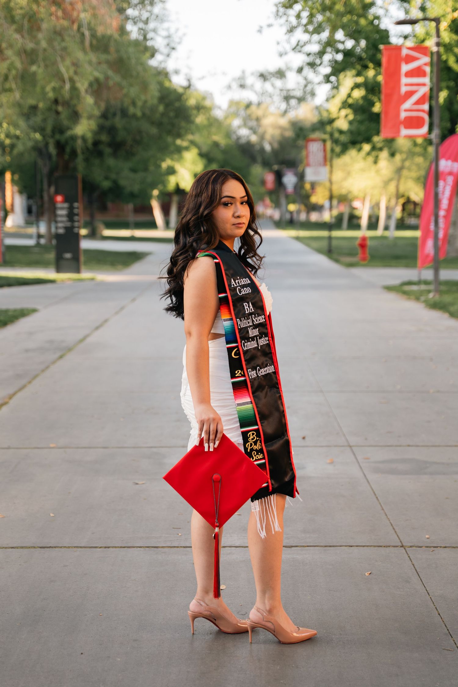Graduate in white dress and graduation stole poses on campus pathway.