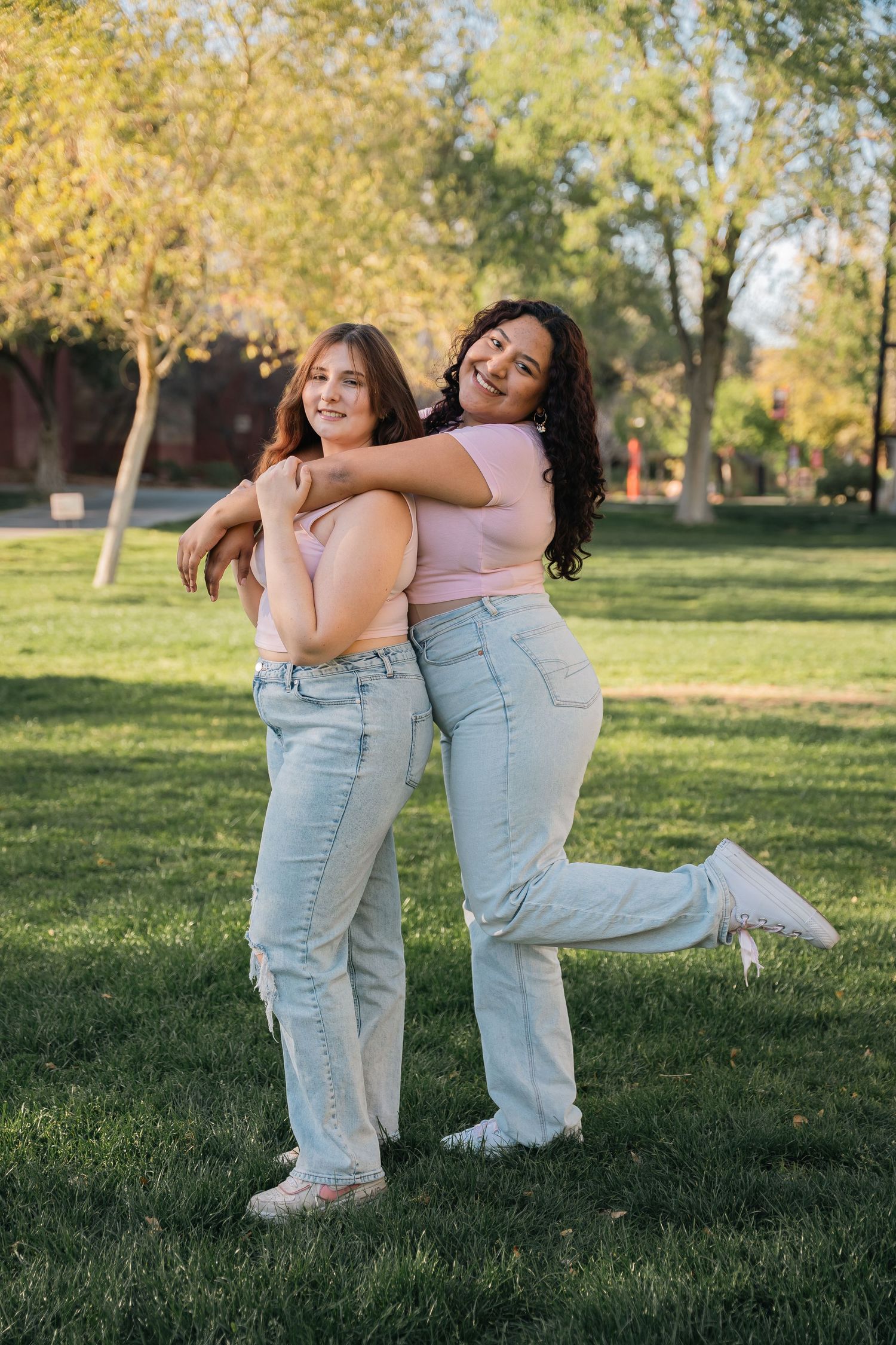Friends in pink tops and jeans embrace playfully while posing on grass with autumn trees behind them.