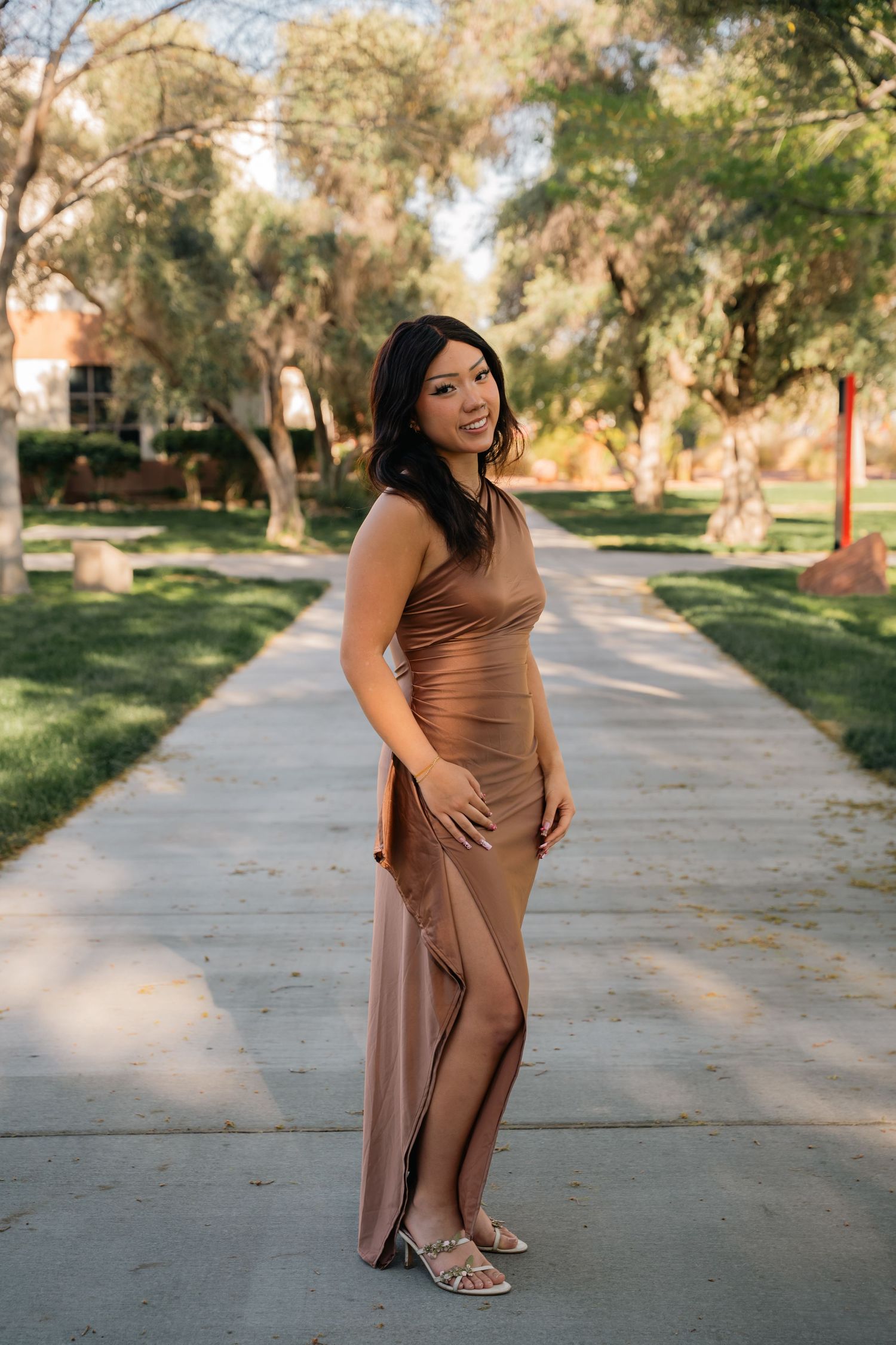Woman wearing elegant brown gown poses on walkway surrounded by lush trees.