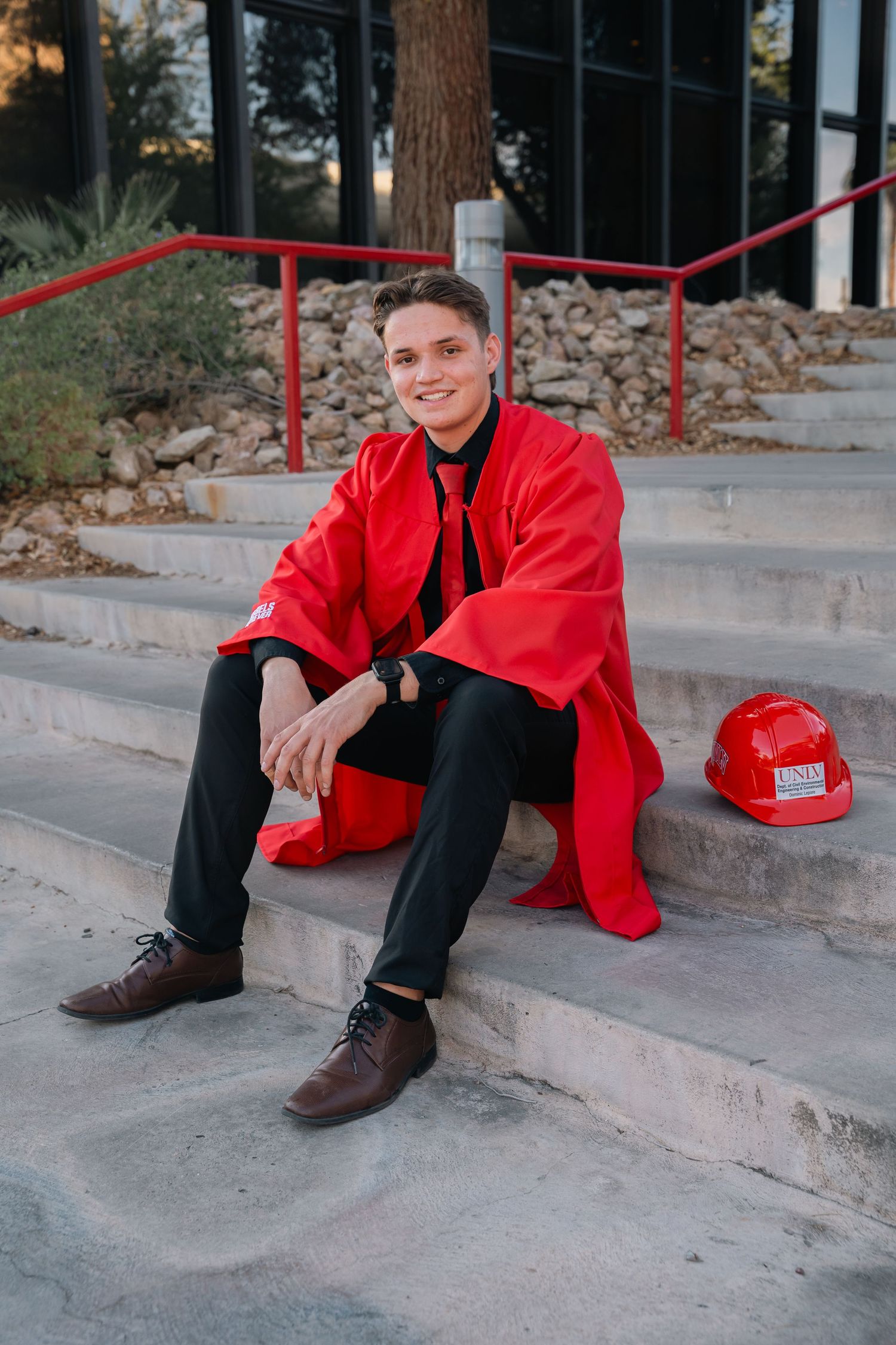Graduate in red graduation gown sits casually on concrete steps outdoors.