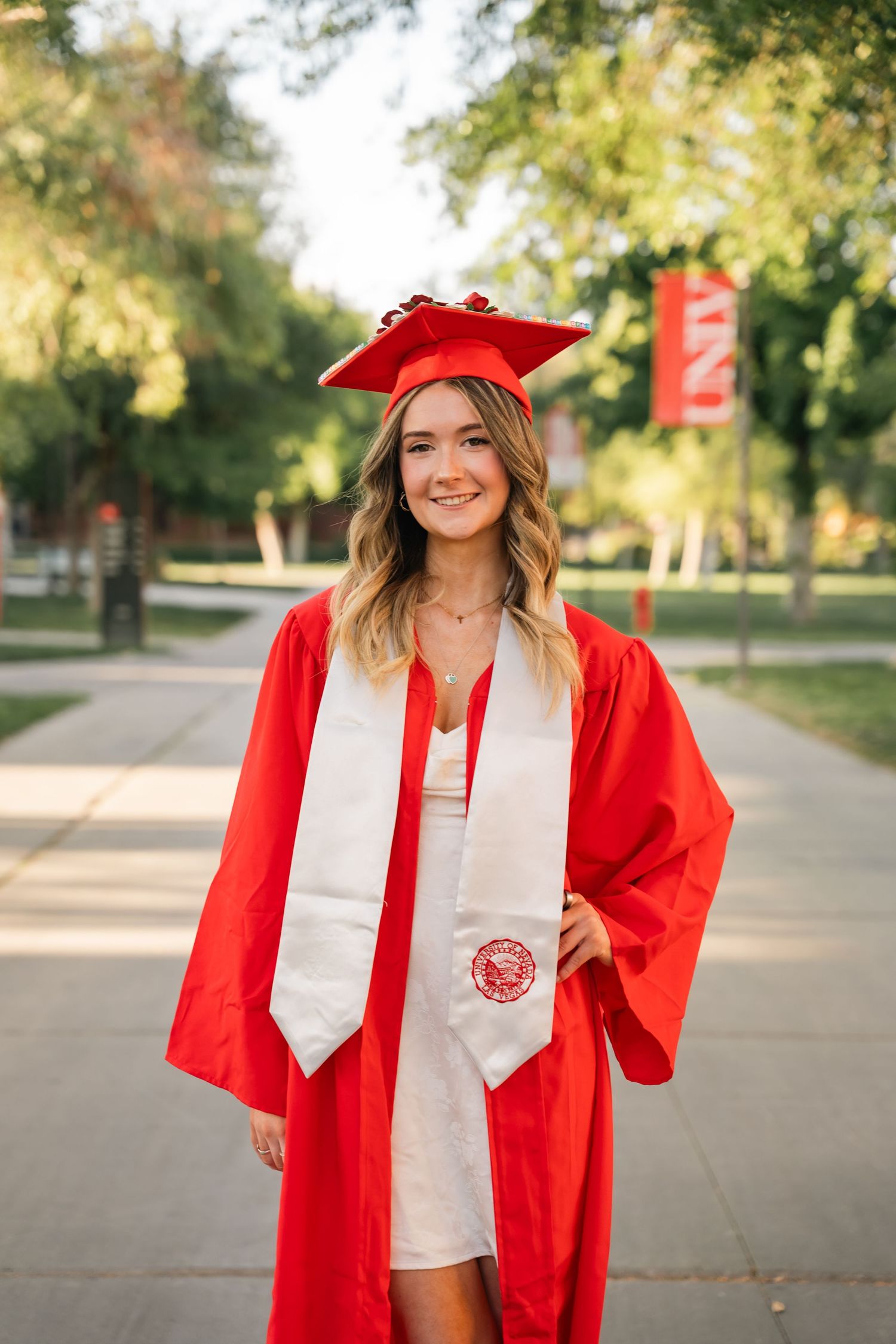 Graduate wearing red cap and gown with white stole stands on campus pathway by UNLV banner.