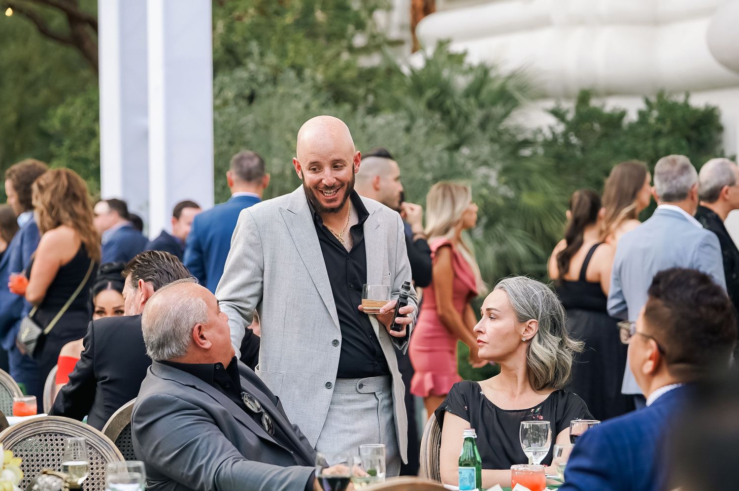 Outdoor wedding reception cocktail hour with guests mingling under white tent columns.