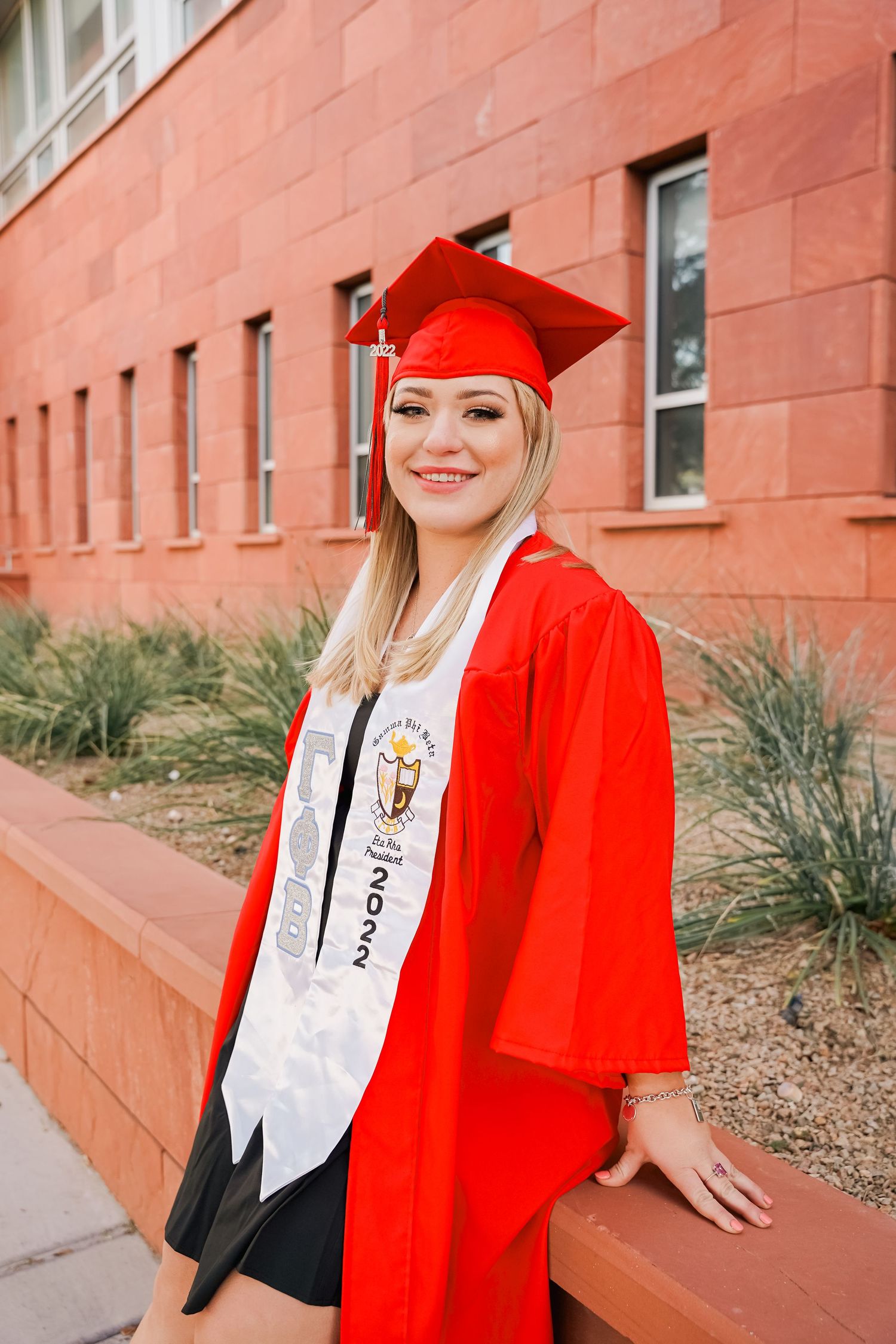 Graduate in red academic regalia smiles while leaning against terracotta colored wall.