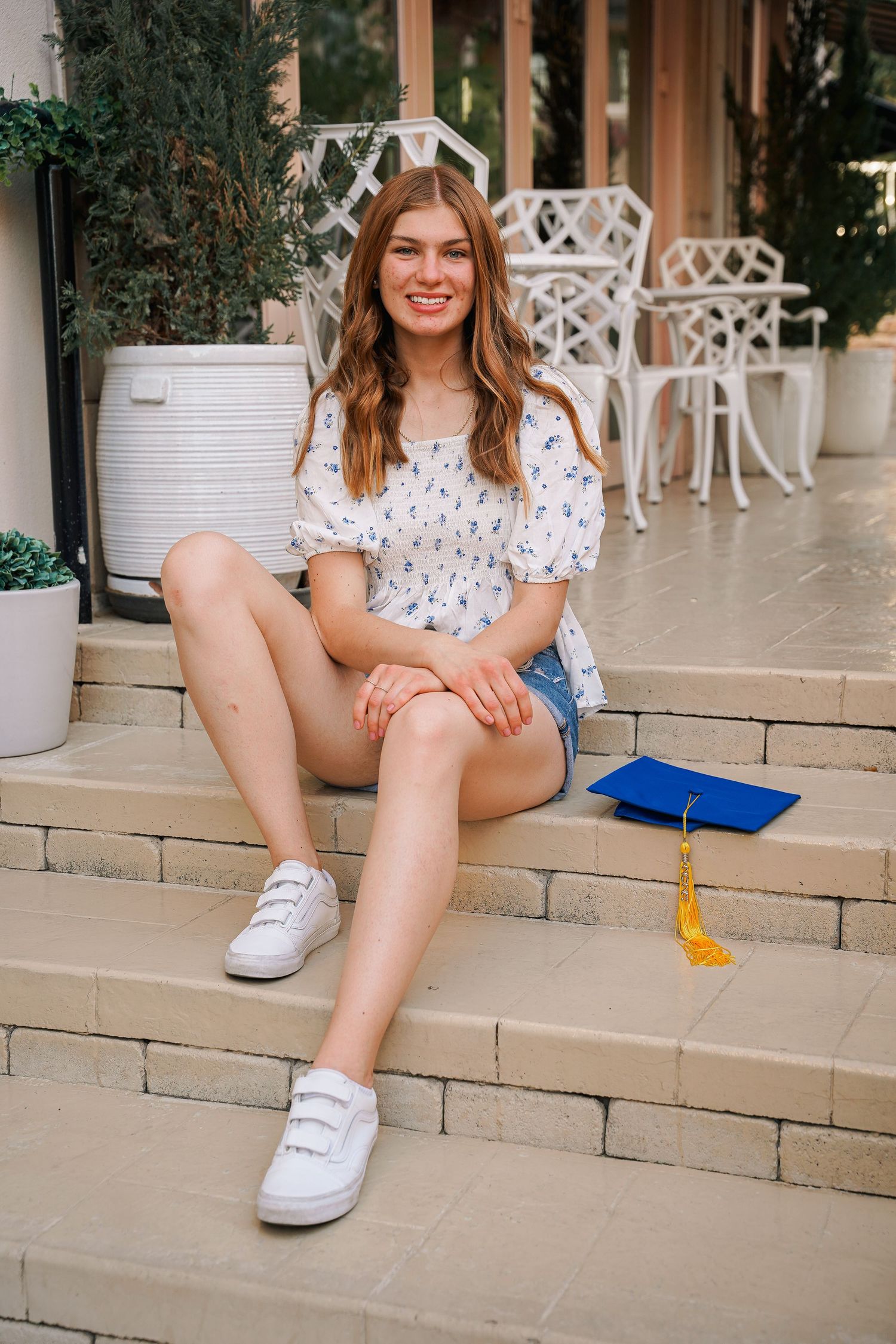 Graduate in casual summer outfit sits on steps with graduation cap nearby.