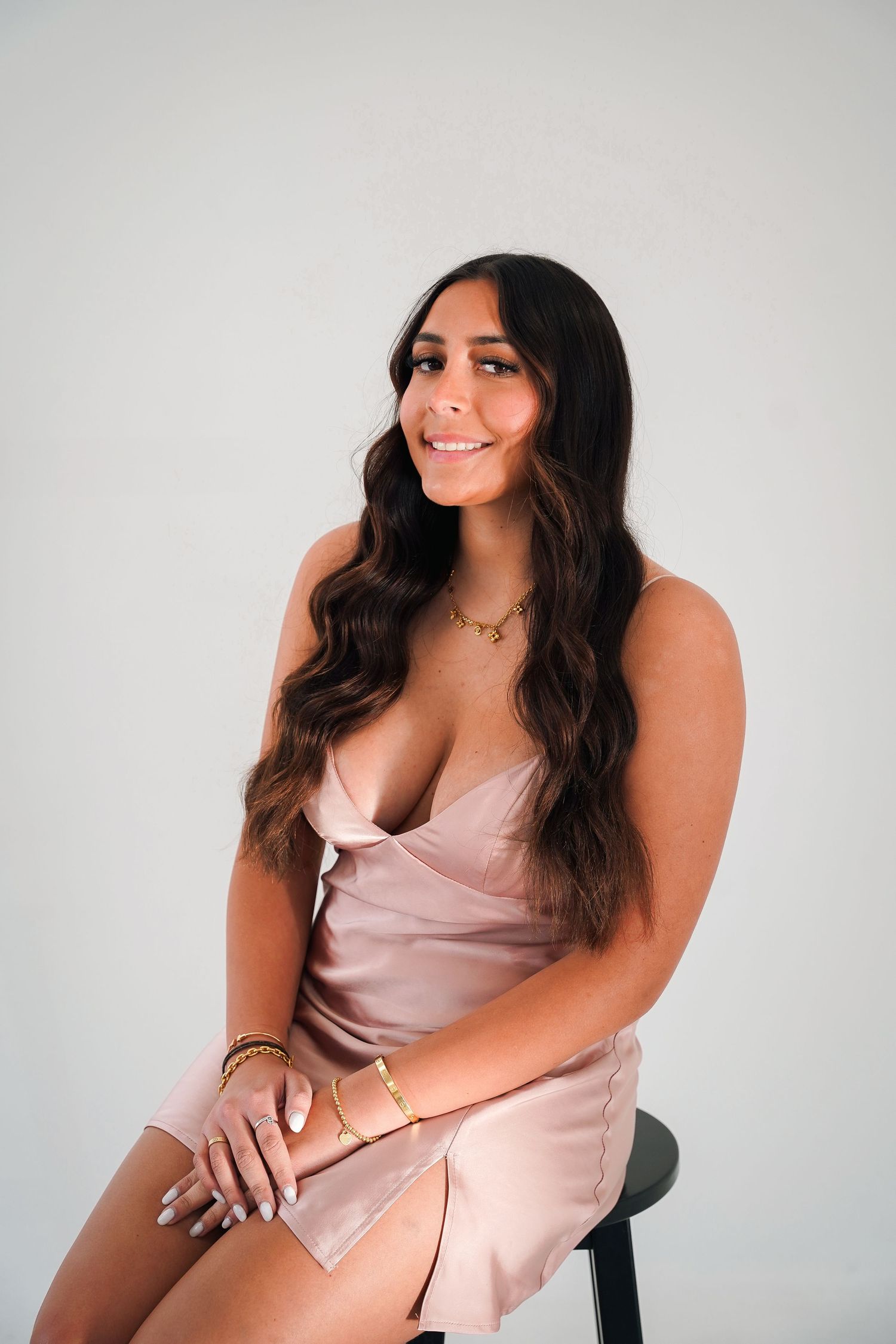 Woman in pink satin dress poses on chair against white studio backdrop.