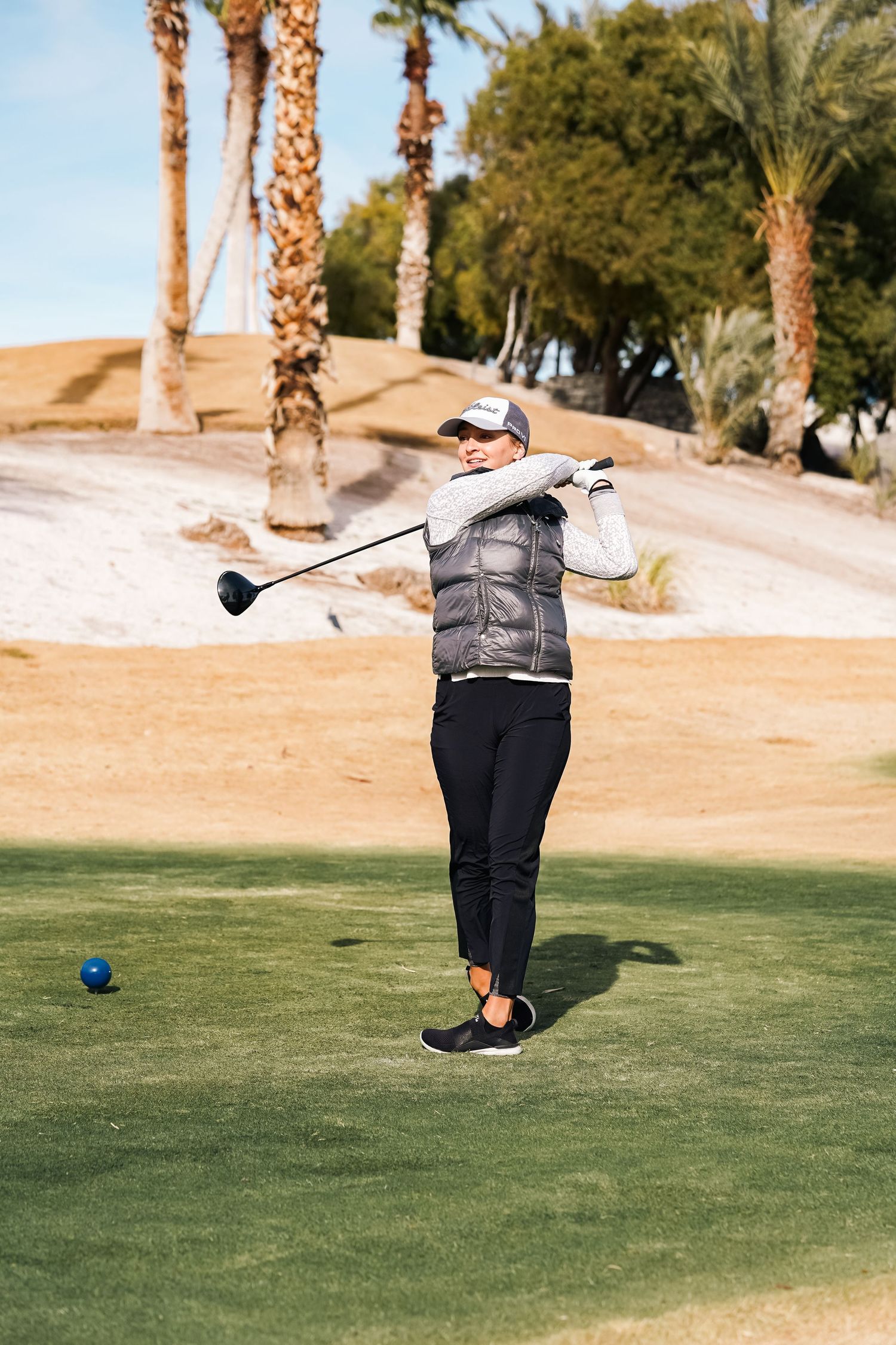 Golfer in a grey vest taking a backswing on a desert golf course lined with palm trees.