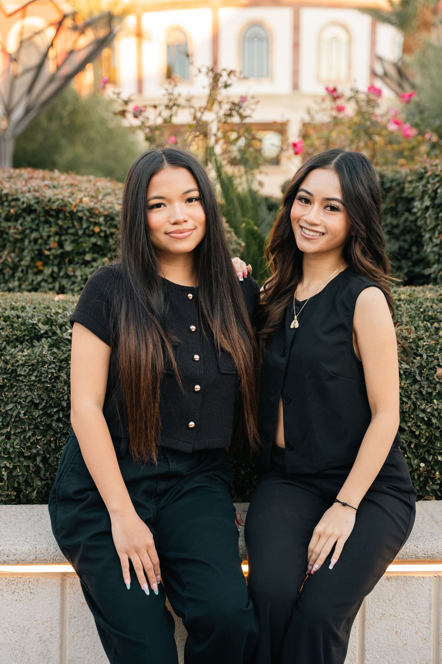 Two friends in matching black outfits sitting together on stone ledge with garden backdrop.