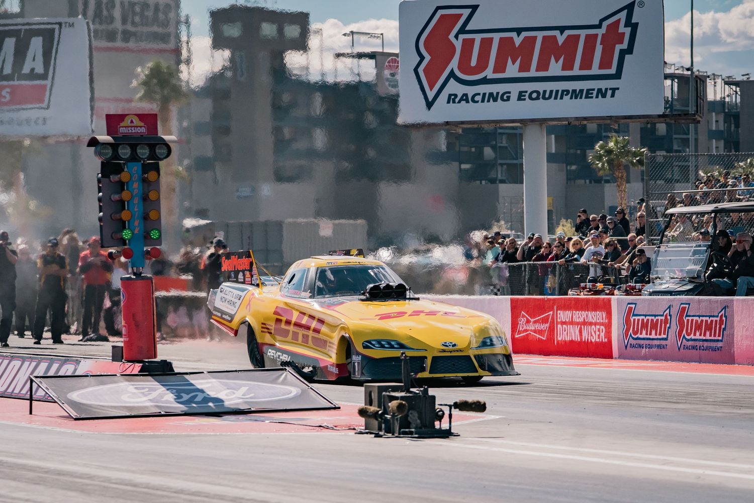 Yellow drag racing car launching from the starting line with timing equipment visible.