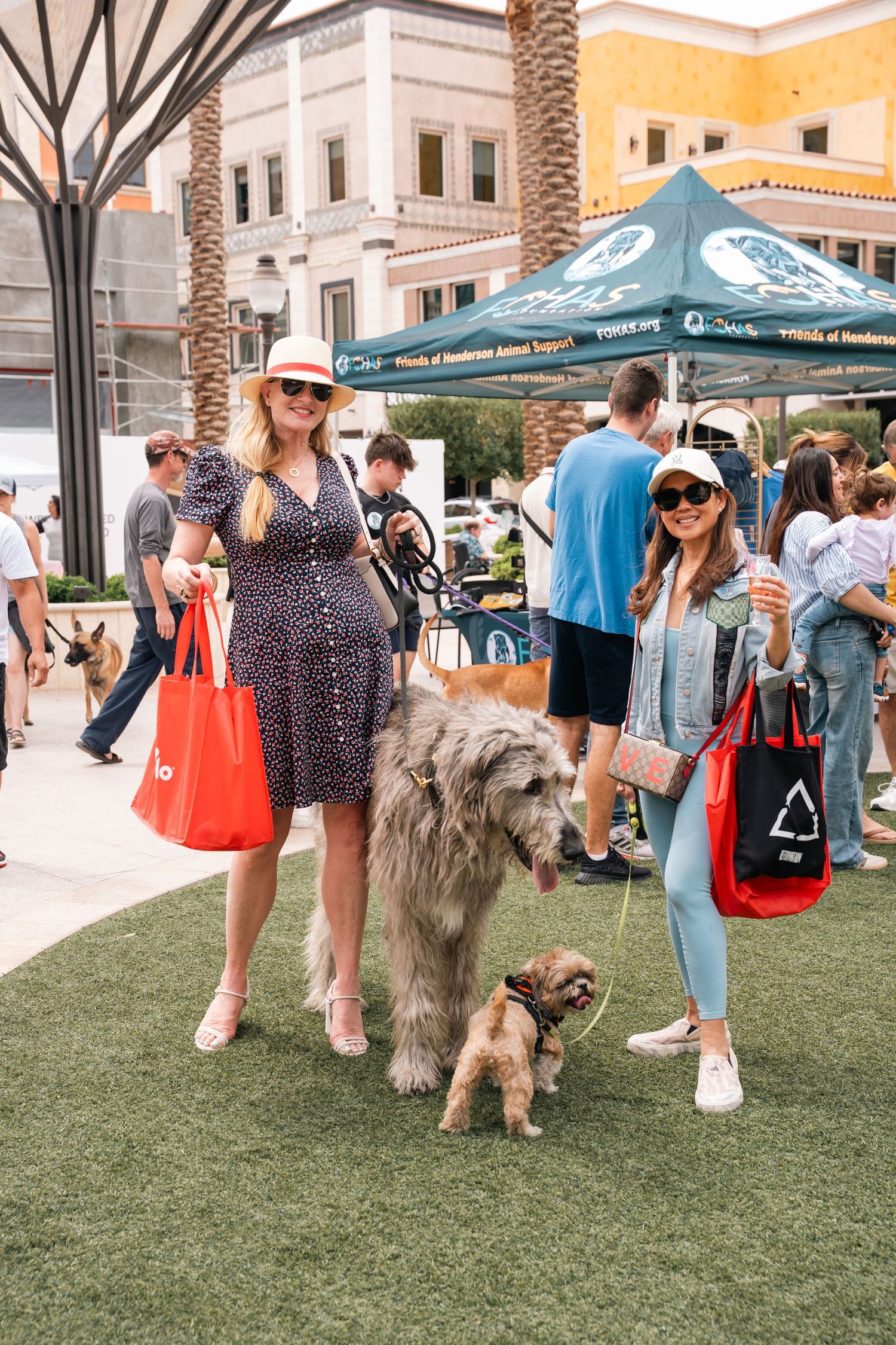 Two dogs and their owners posing at outdoor community event with palm trees in background.