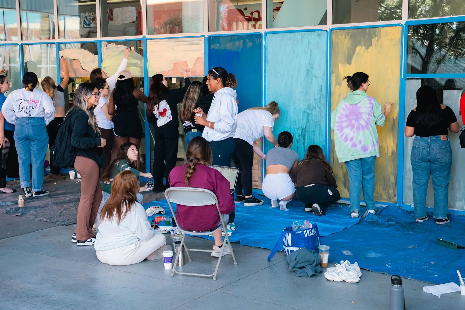 Students painting panels on blue tarp with supplies and backpacks scattered around.