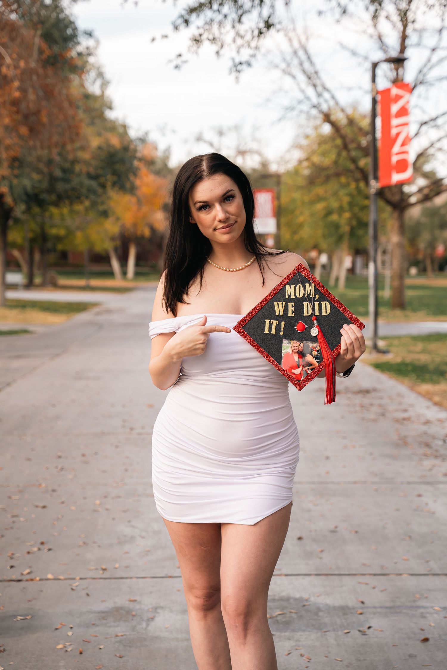 Graduate in white dress holding decorated mortarboard cap outdoors.