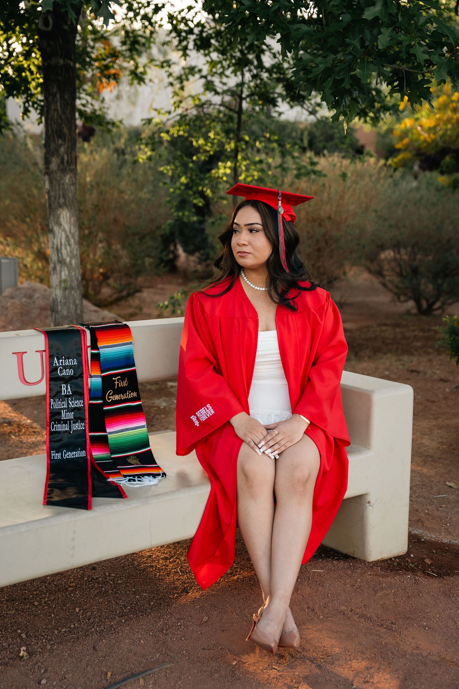 Graduate in red regalia sits on stone bench with graduation stole and traditional serape.
