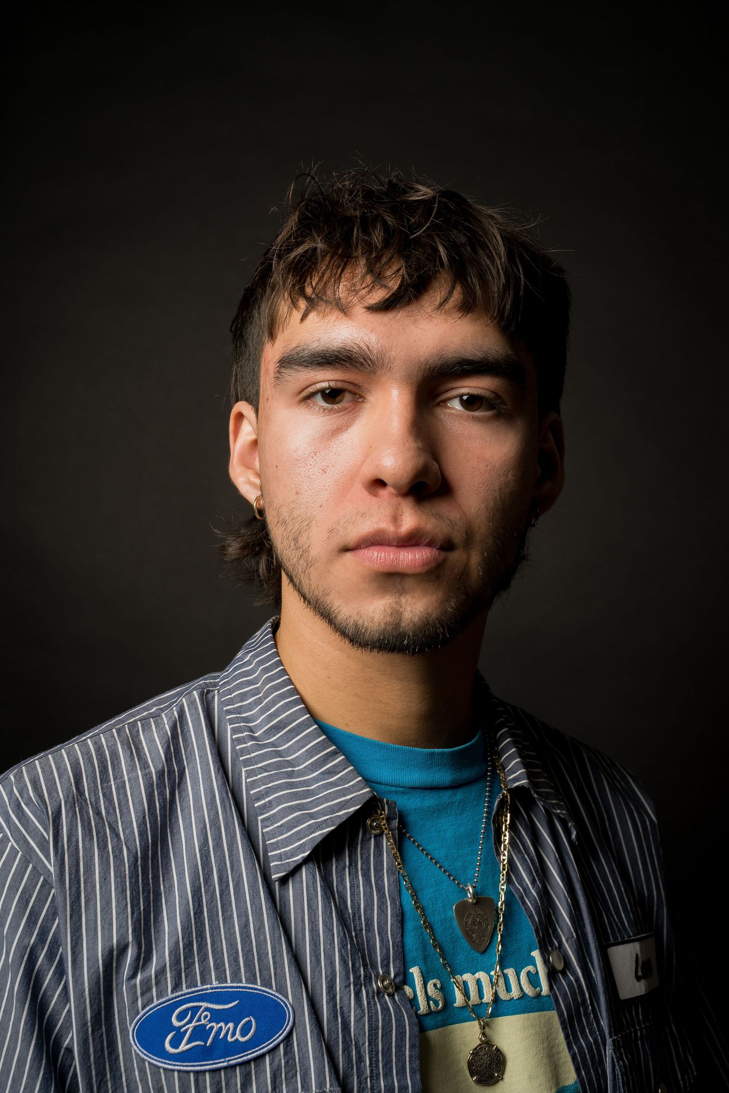 Monaco Media. Close-up dramatic portrait of a young man wearing blue shirt and striped button-up in low lighting.