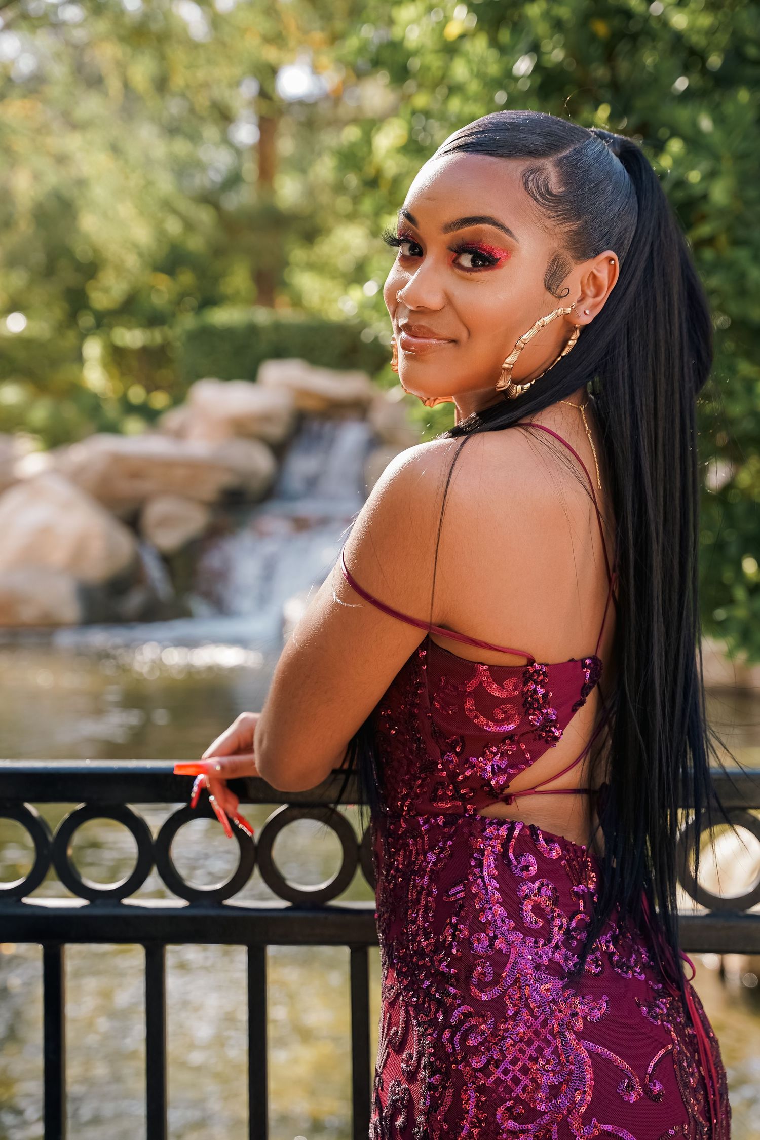 A person in a sparkling burgundy formal dress poses by a waterfall with an ornate metal railing.