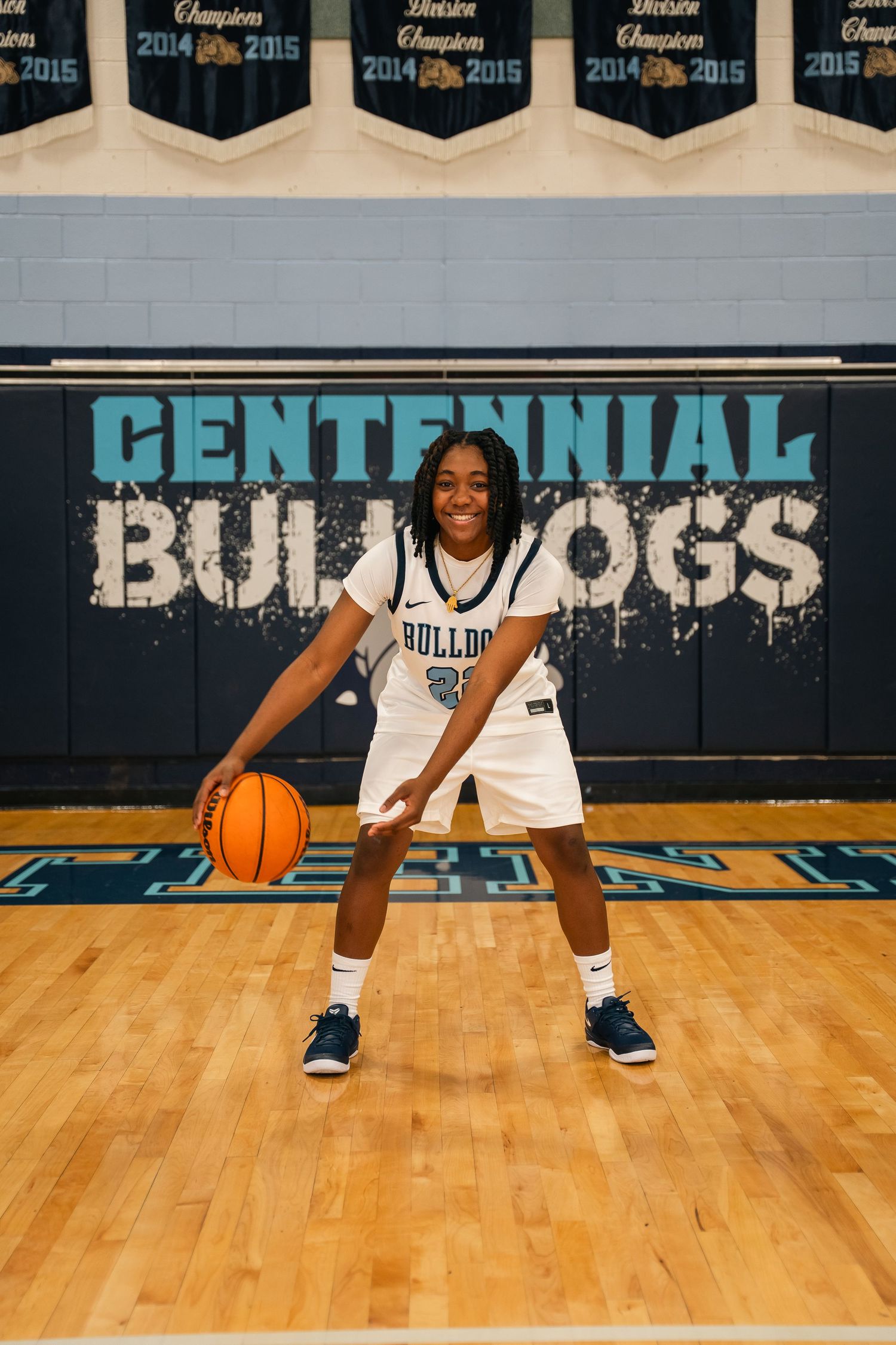 Basketball player in white Bulldogs uniform dribbling on wooden court with championship banners.