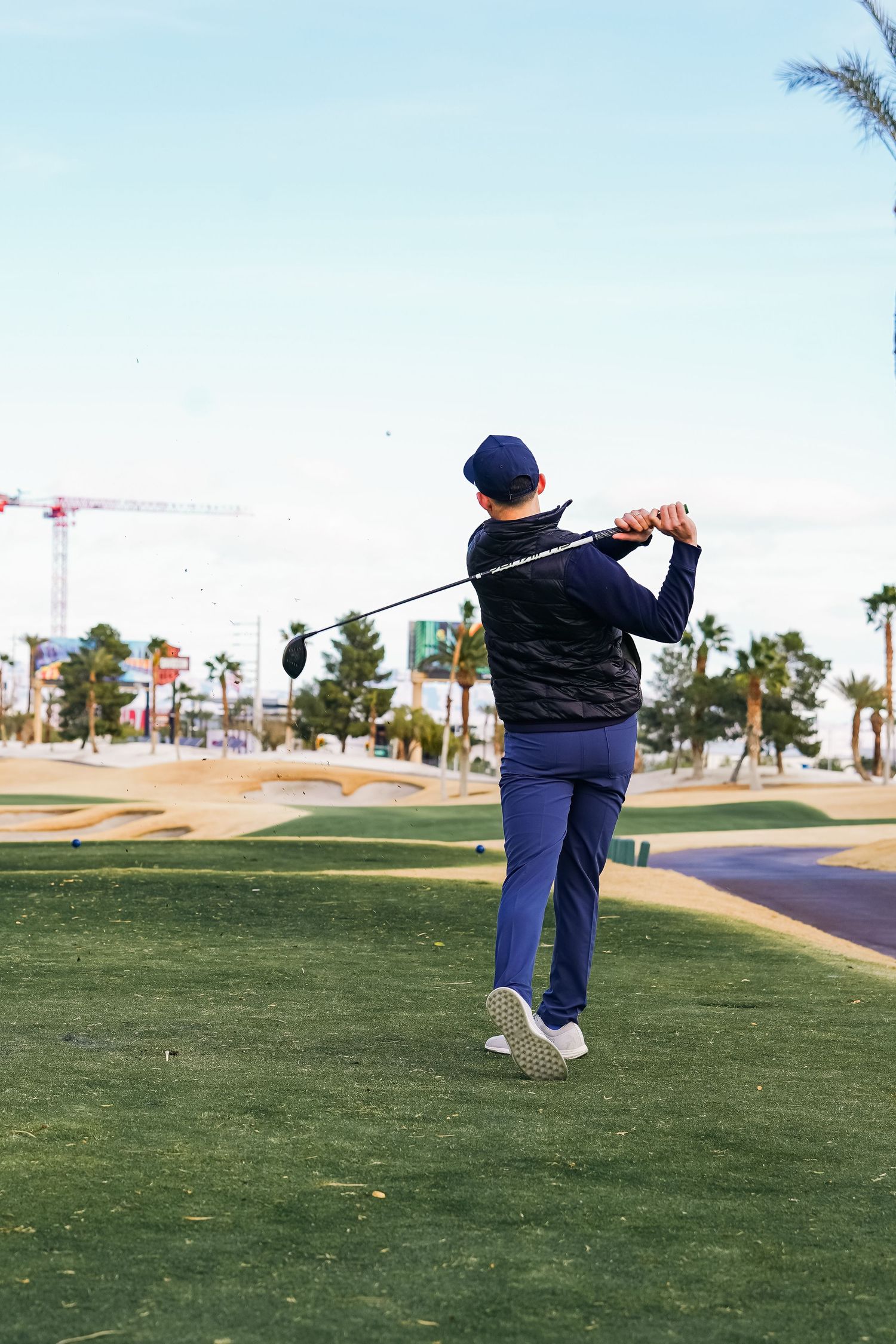 Golfer follows through on swing at desert golf course with palm trees and buildings in view.