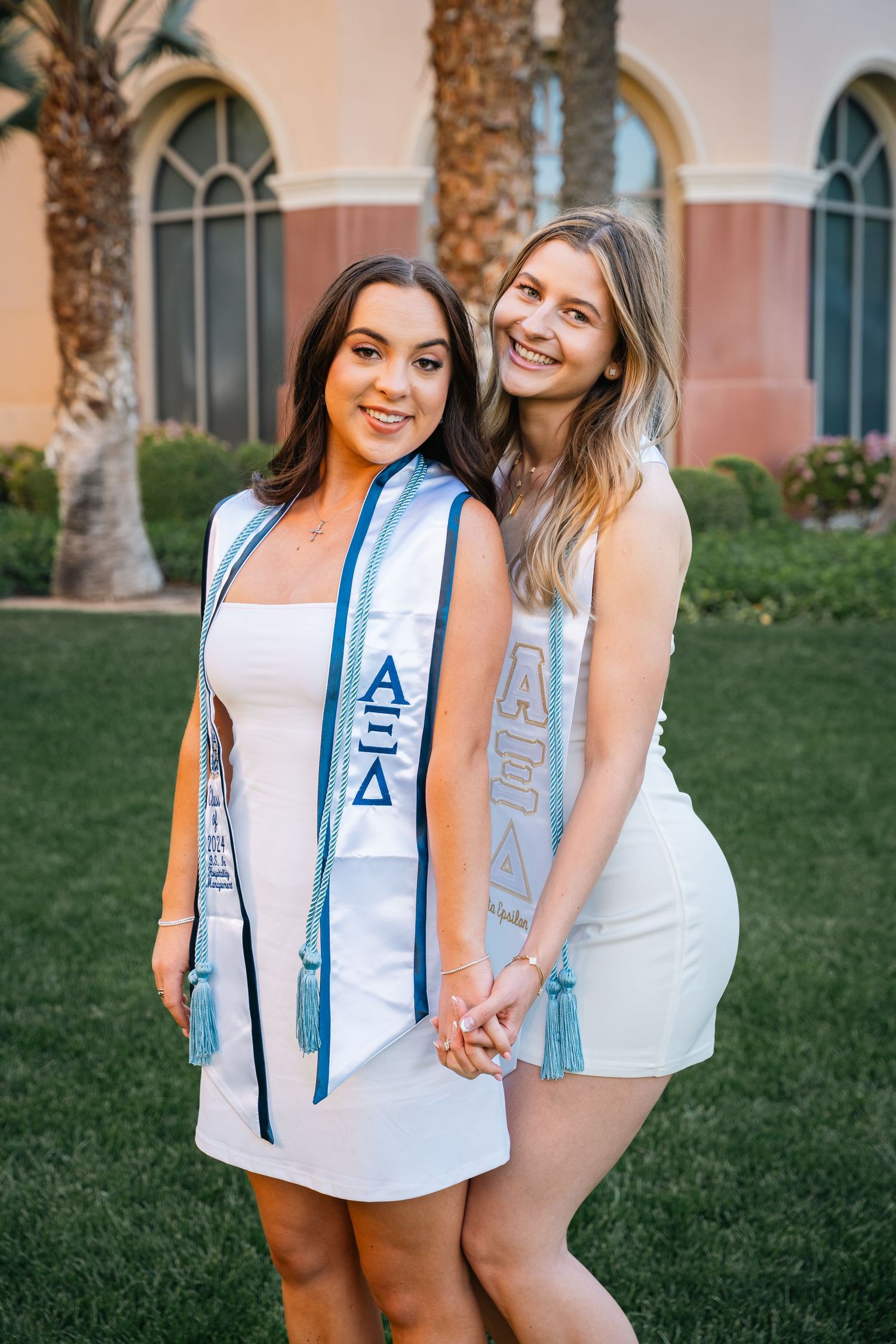 Two sorority sisters in white dresses and light blue stoles pose together on grass.