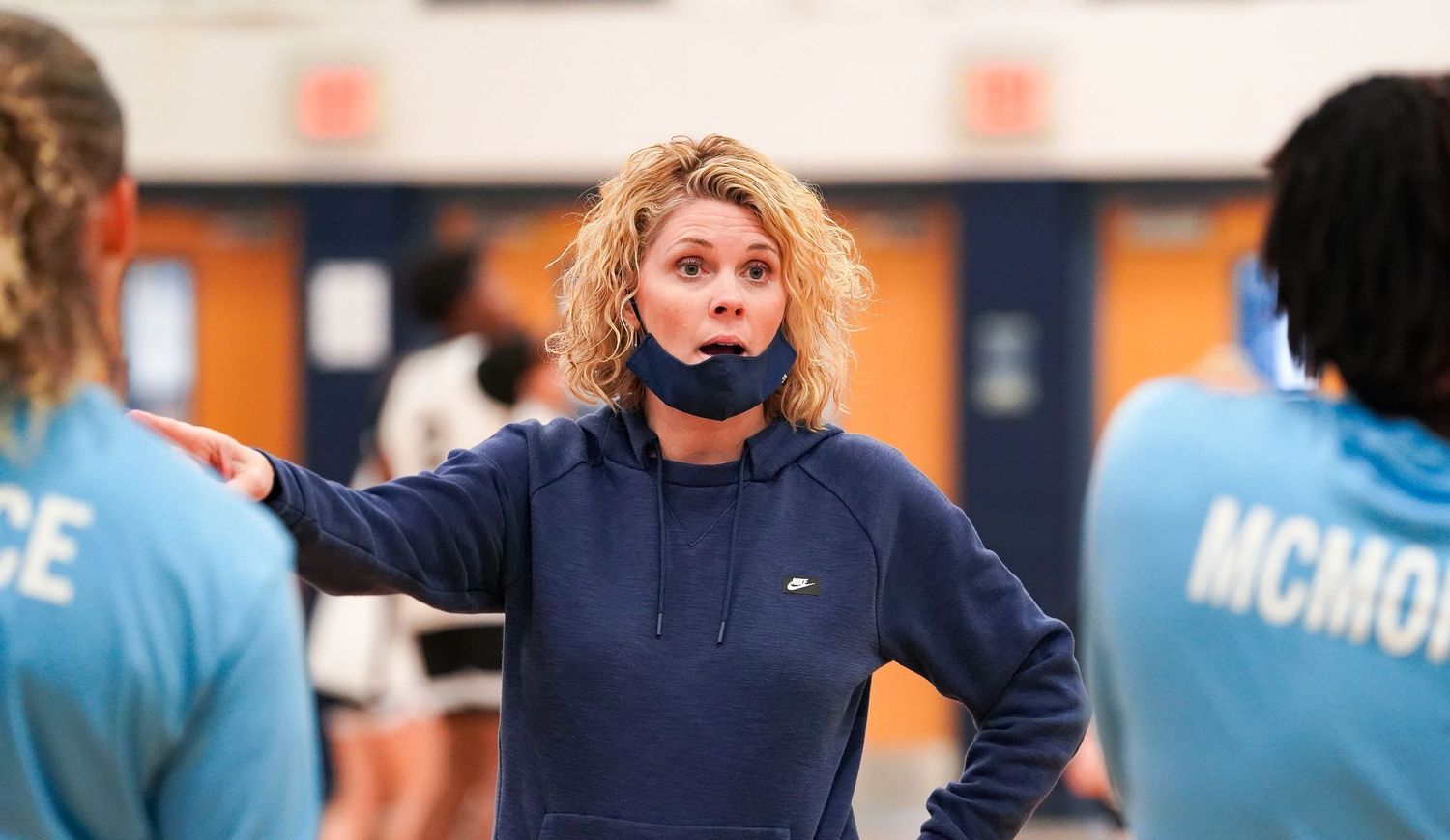 Coach wearing navy hoodie and face mask gives instructions during basketball practice.