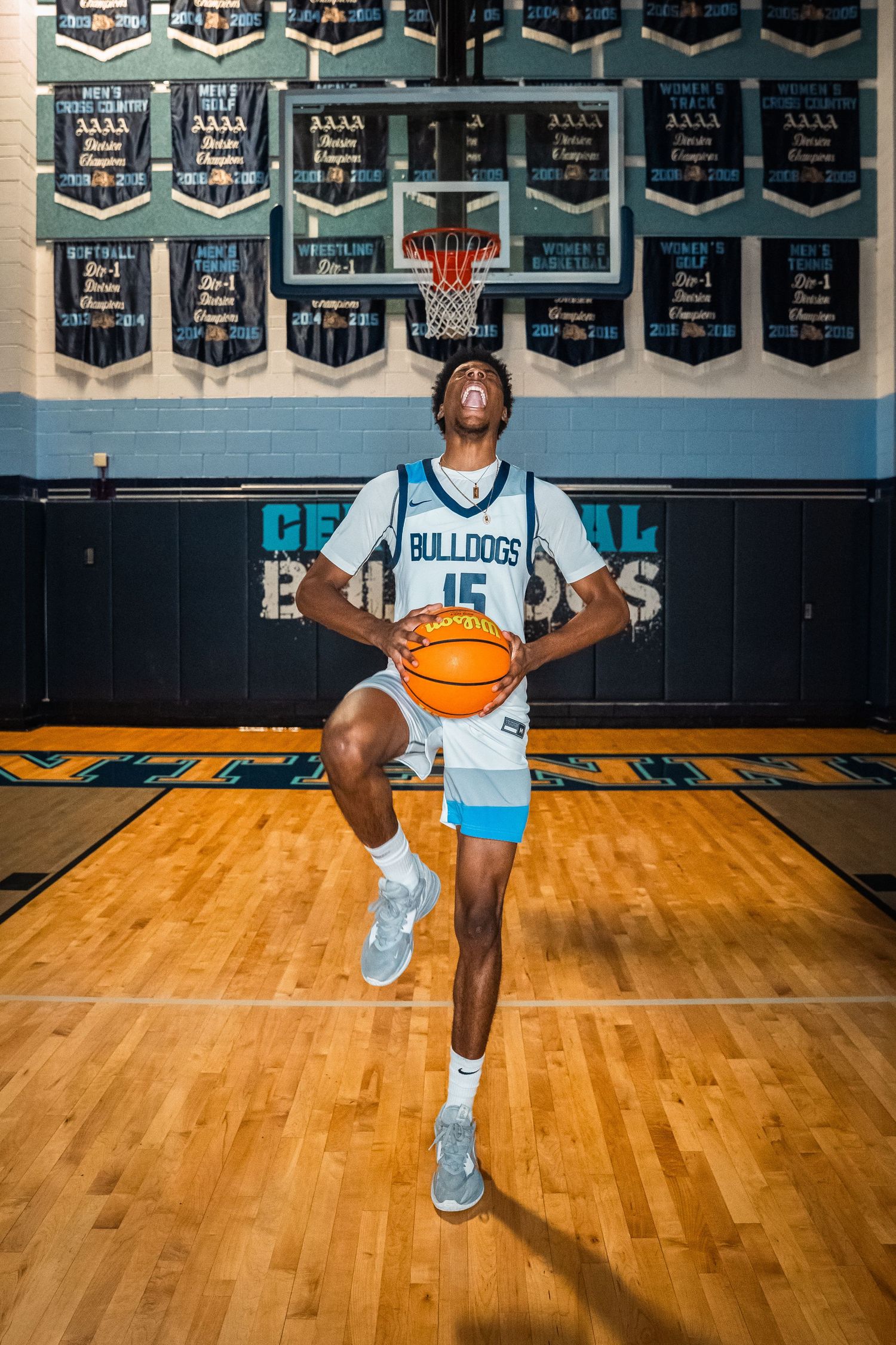 Basketball player in light uniform poses with ball on wooden court under championship banners.