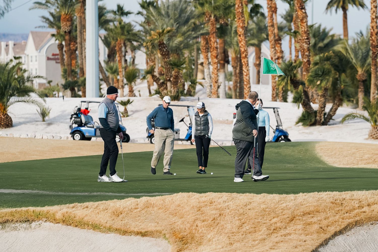 Group of golfers gathered on green desert course with palm trees and resort buildings.