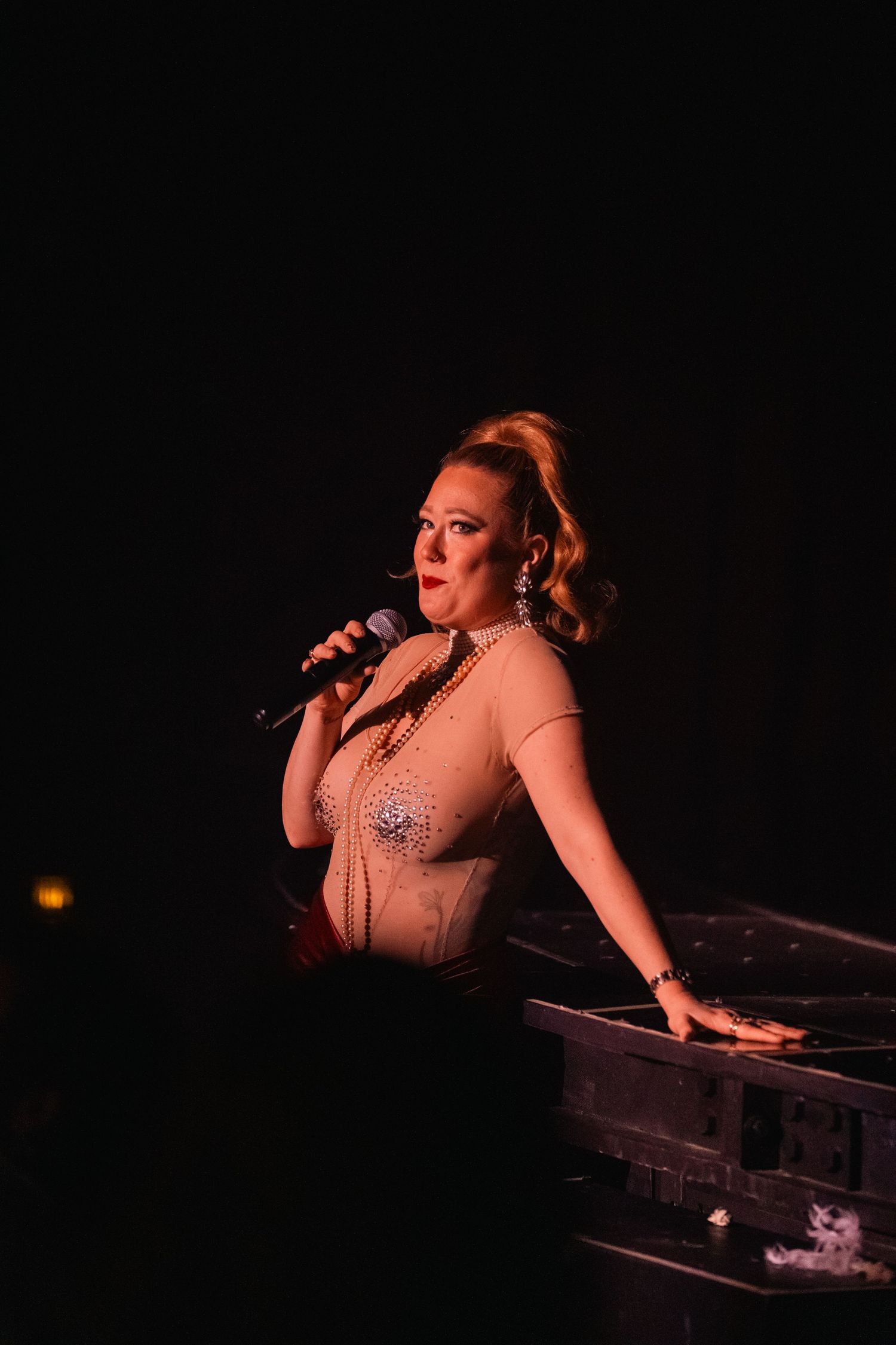 Performer in a sparkly beige corset singing on stage in dramatic lighting.