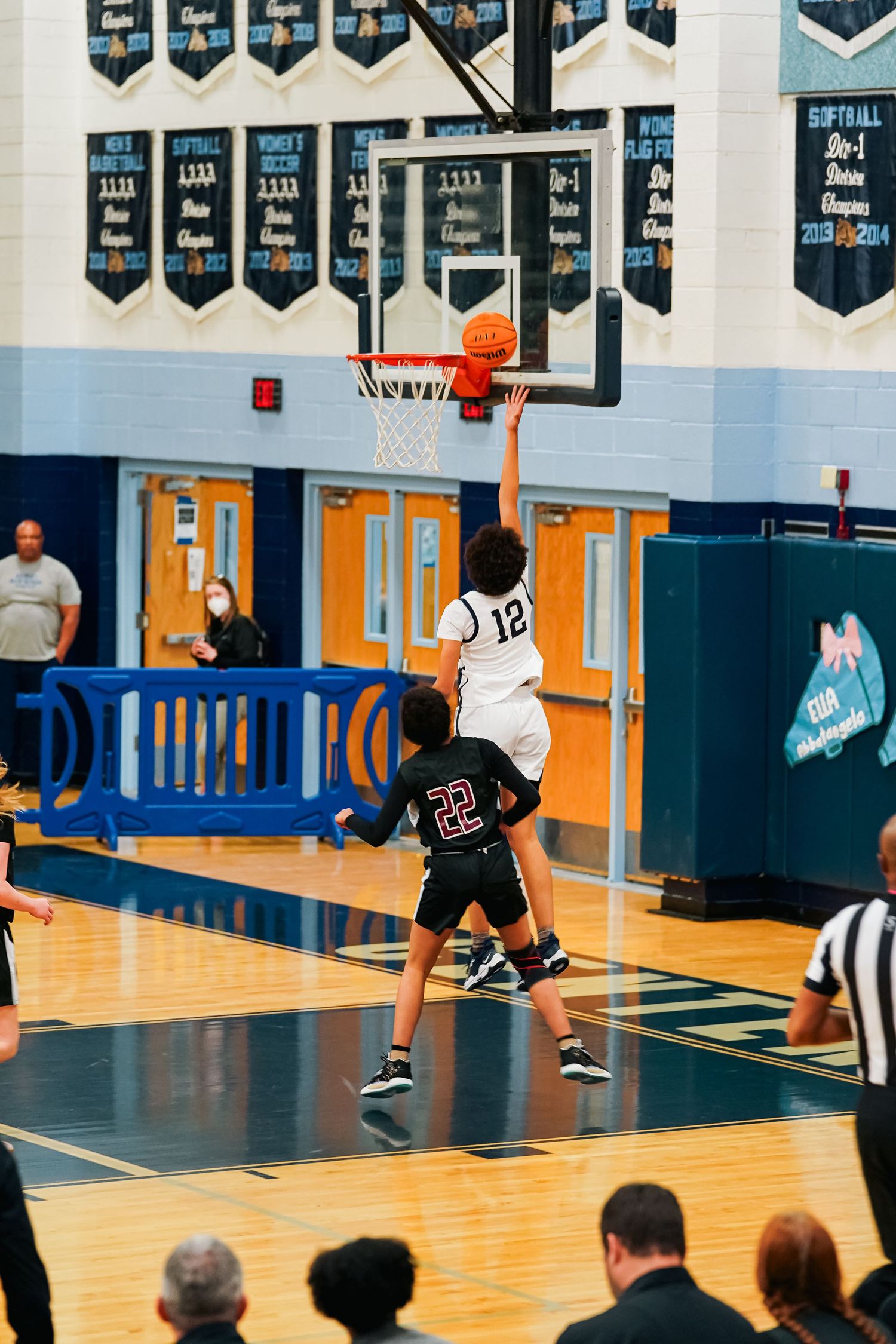 Basketball game action shot with players and referees on court with championship banners visible.