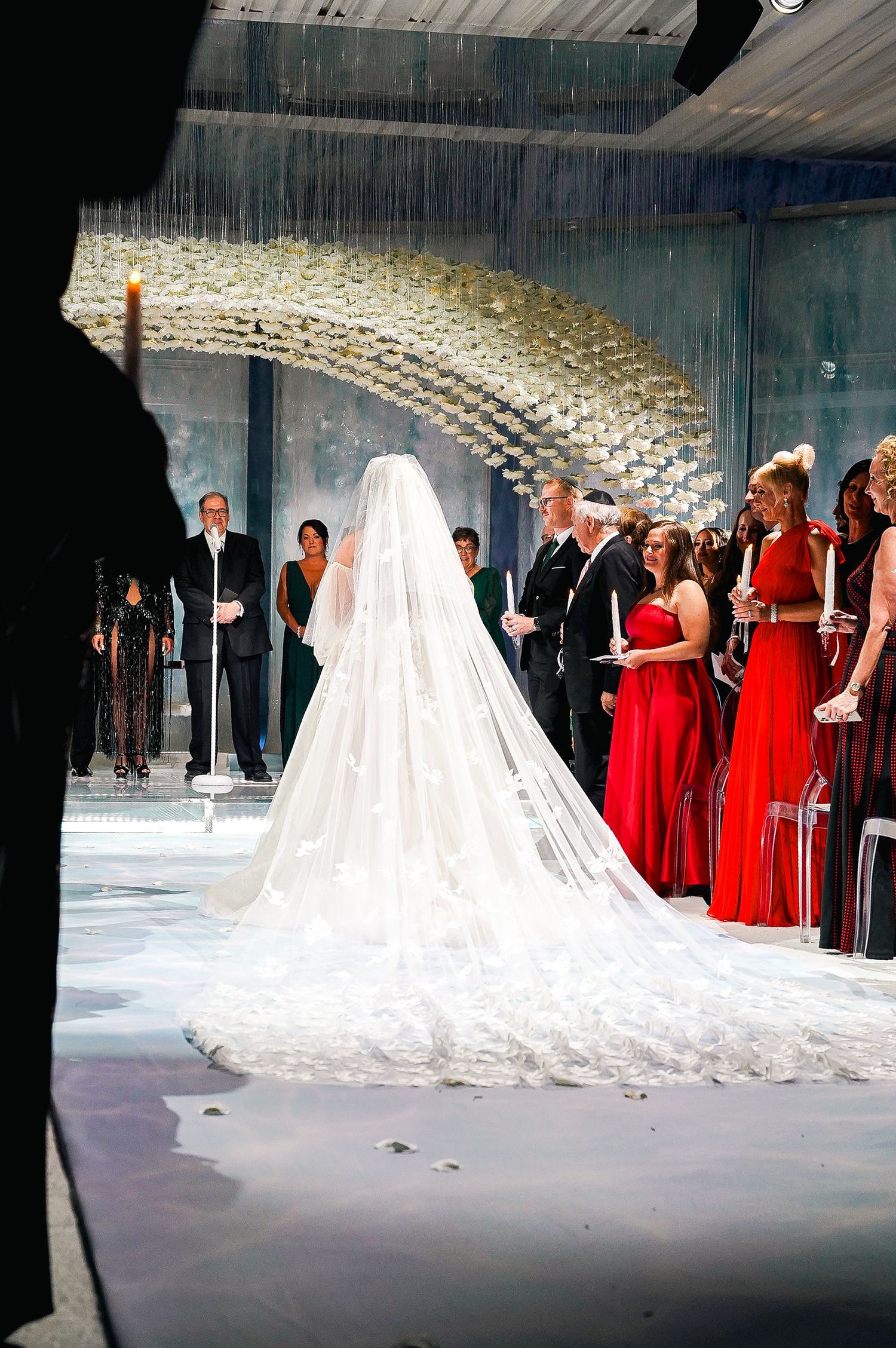 Back view of bride in cathedral length veil with red-dressed bridesmaids at ceremony.