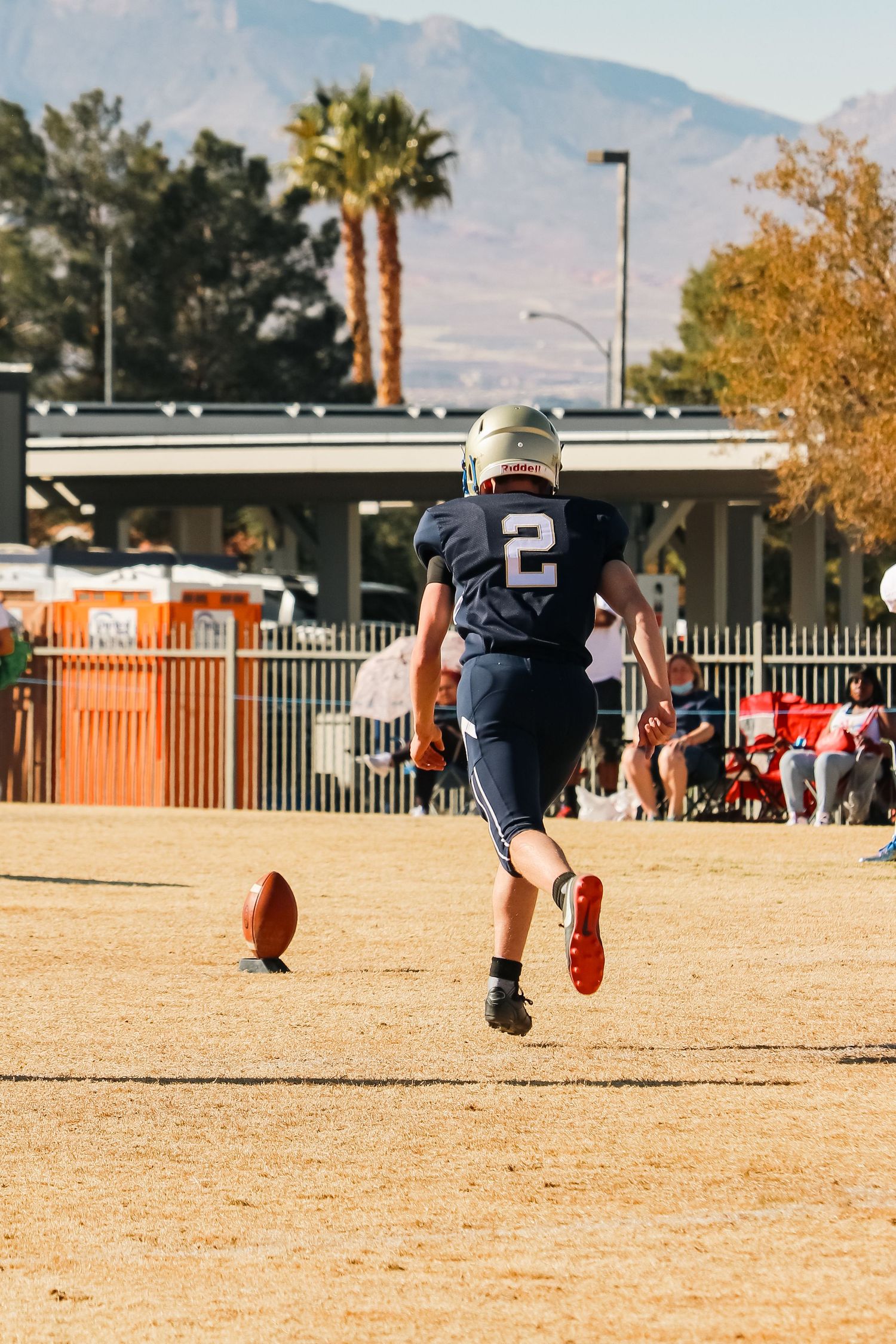 Football player in navy uniform running towards football on field with mountains in background.