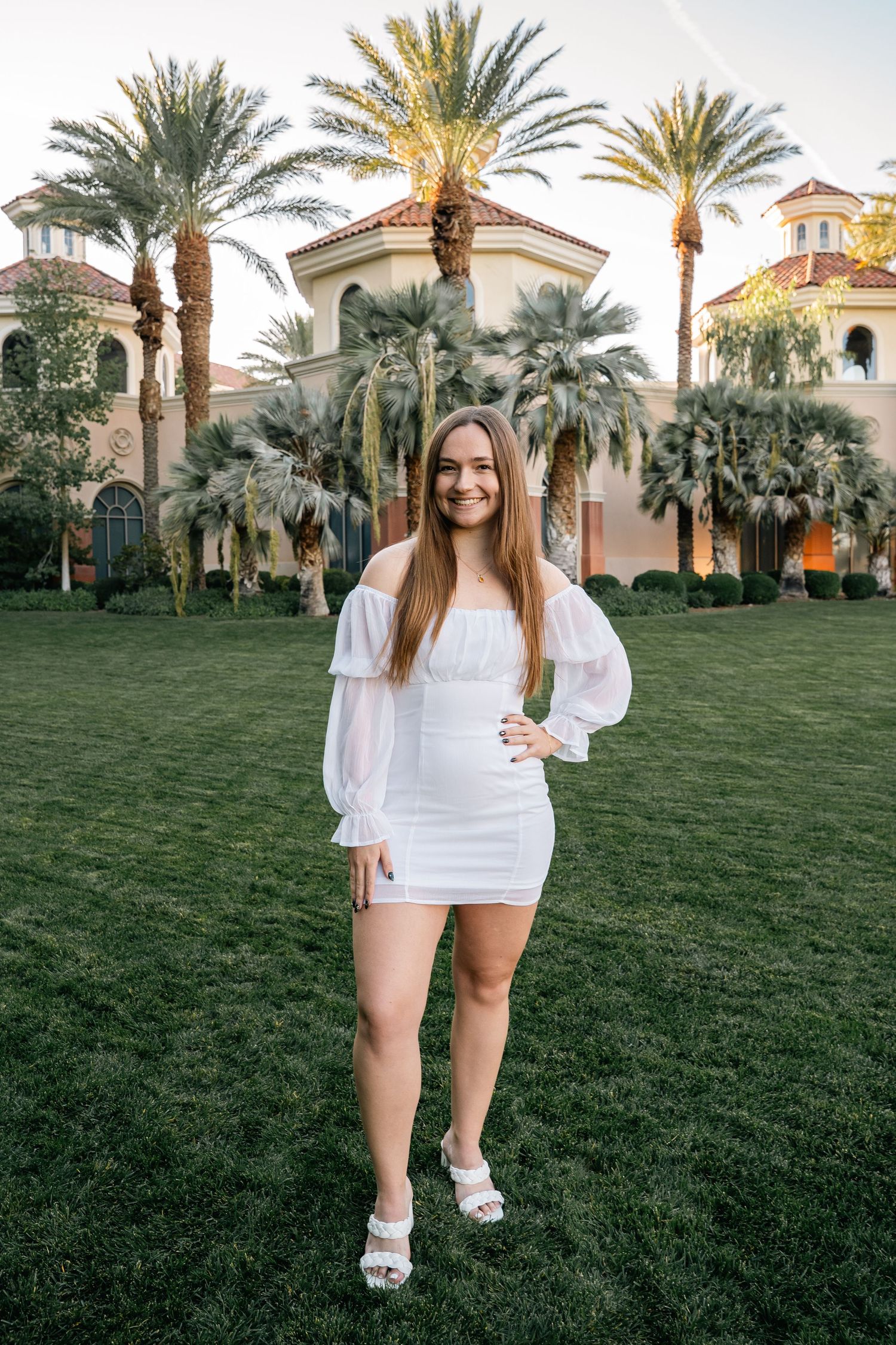 Person in white outfit stands on lawn with palm trees and resort buildings in warm evening light.
