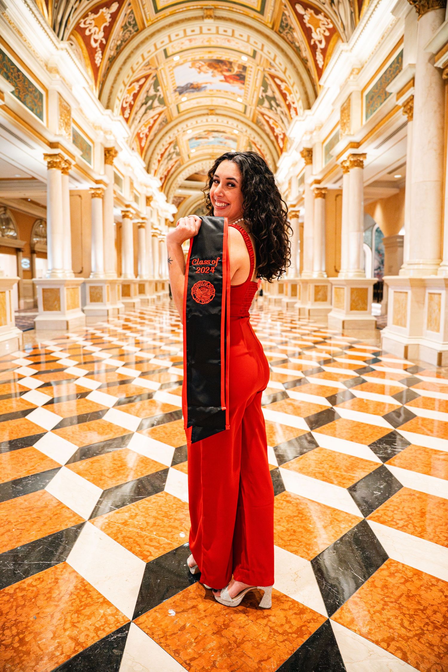 Graduate in red dress posing in ornate hallway with checkerboard marble floor.