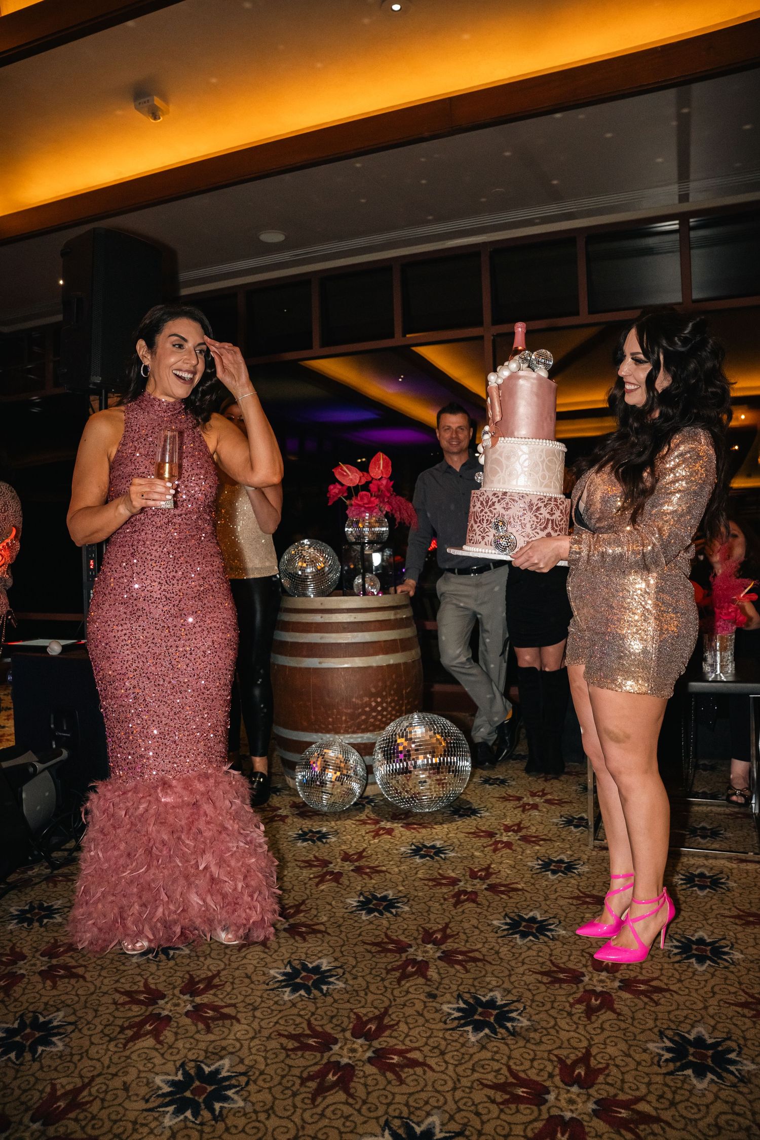 Two guests in sparkly dresses stand by a metallic pink birthday cake at an elegant party venue.