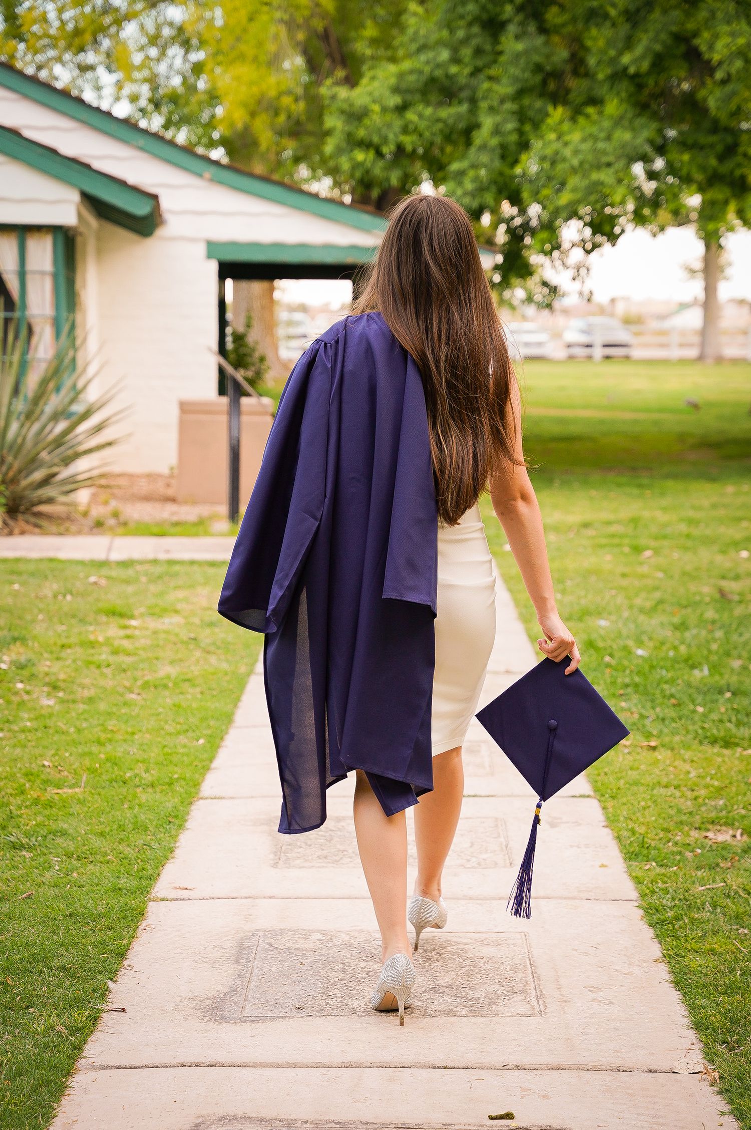 Graduate walks away holding cap while wearing graduation gown on campus pathway.