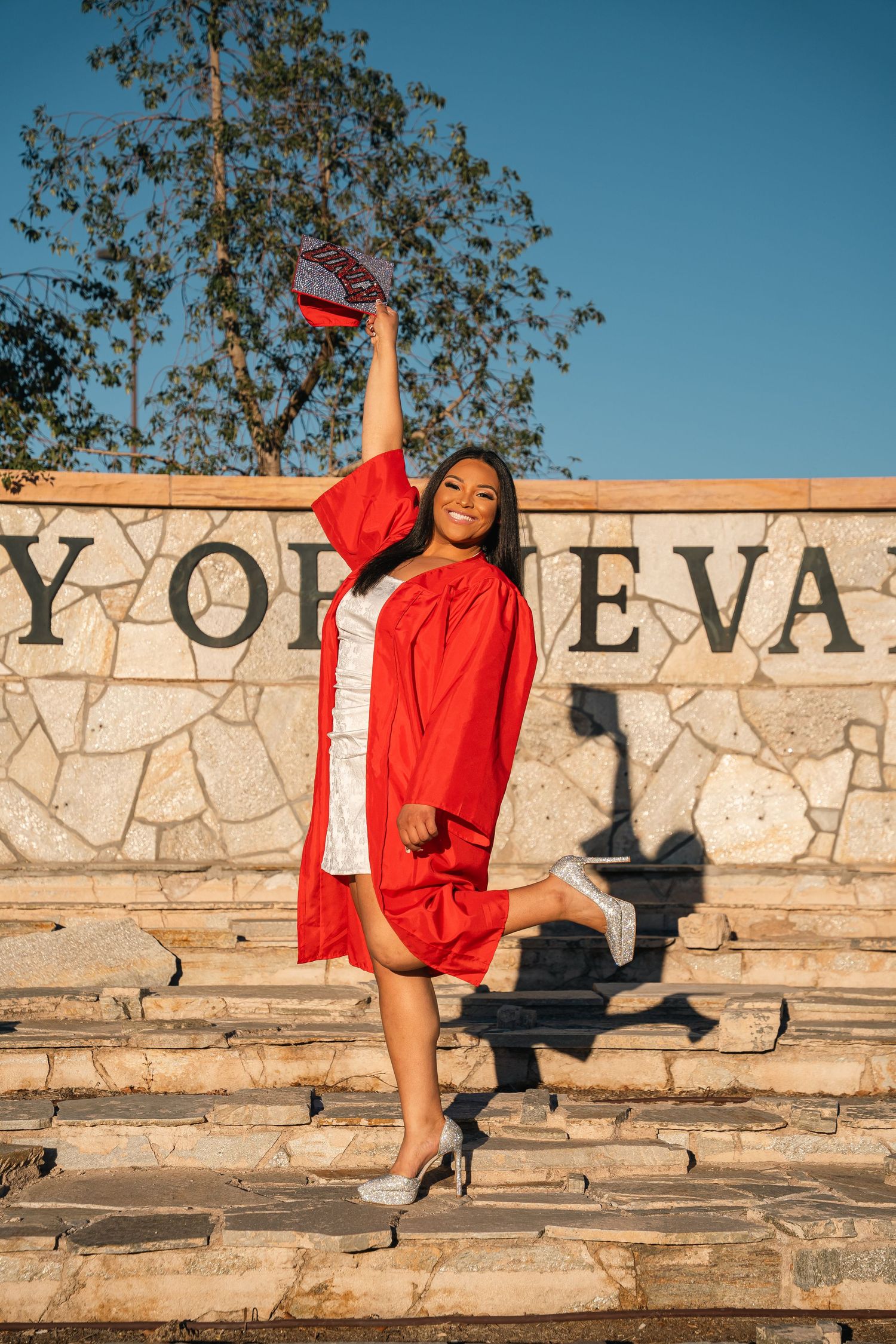 Graduate in red cap and gown poses triumphantly on stone steps with text wall behind.