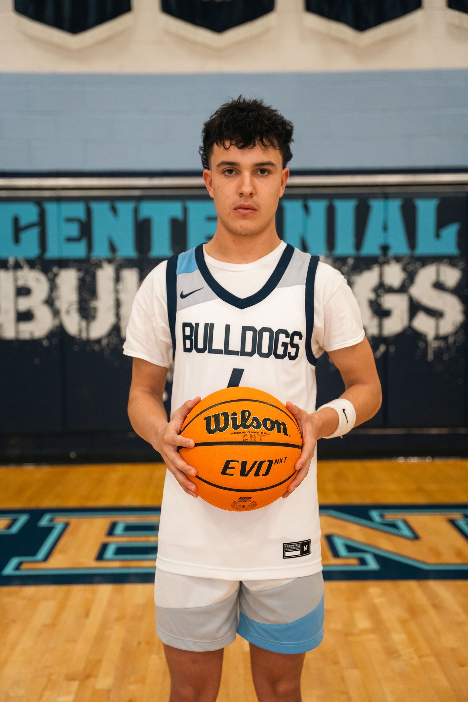 Basketball player in white Bulldogs uniform holding a Wilson basketball on court with championship banners.