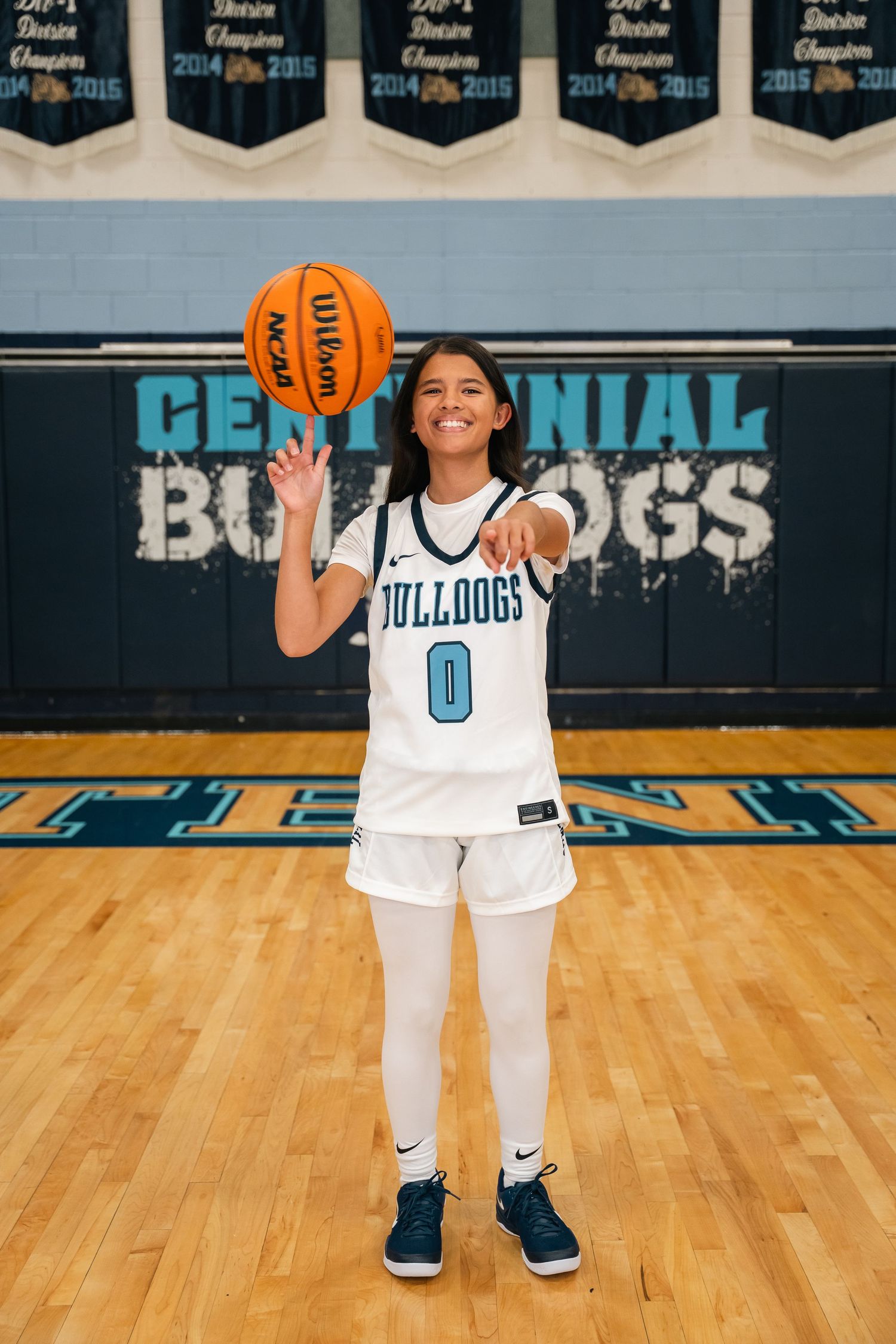 Basketball player in white Bulldogs uniform number 0 holding orange ball on wooden court with championship banners.