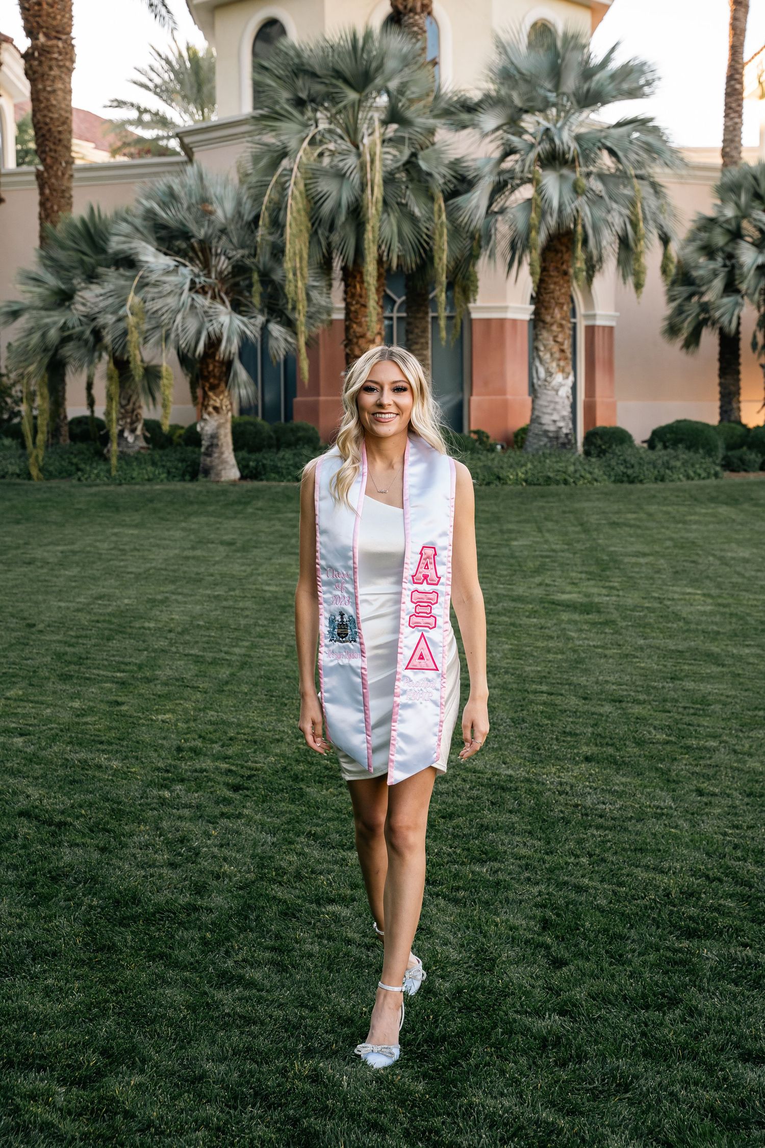 Graduate in white dress wearing AΞΔ stole walks on lawn with Mediterranean architecture in background.