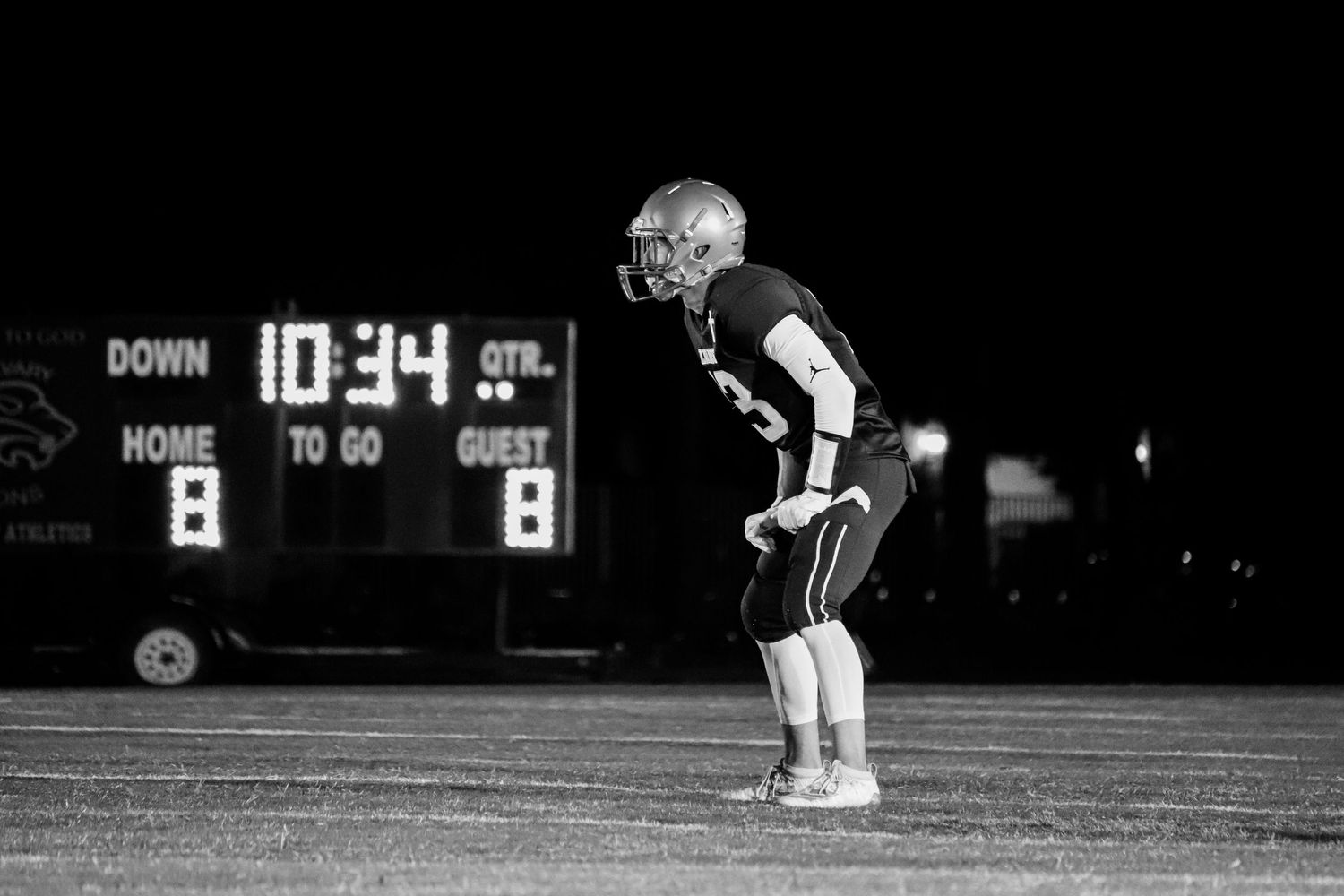 Football player in dark uniform stands ready on nighttime field with scoreboard showing 10:34 in background.
