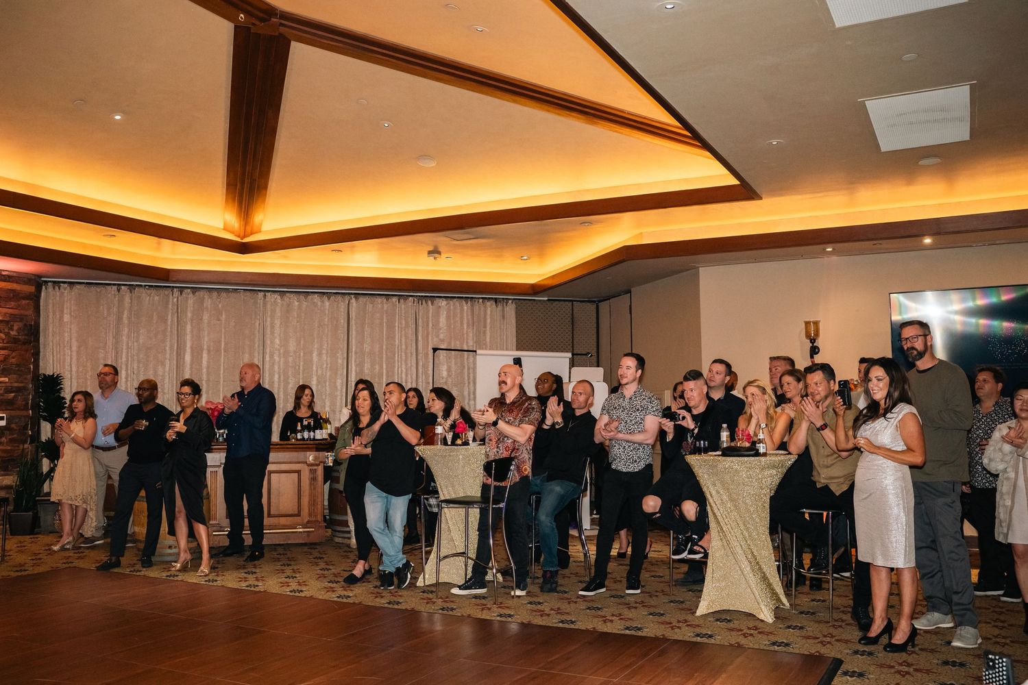 Large group of attendees standing and mingling at an indoor event with warm lighting and exposed ceiling beams.