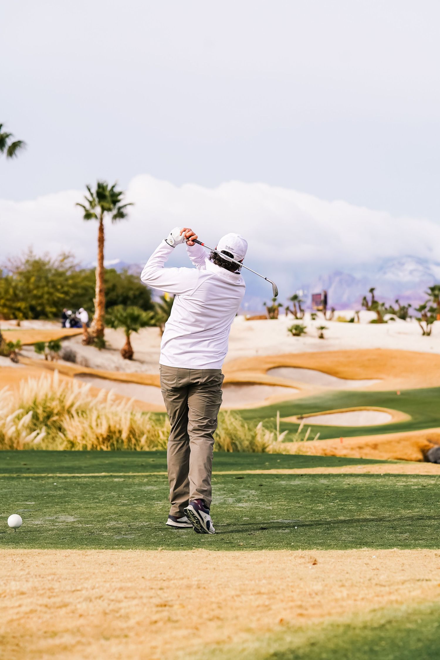 Golfer in white shirt takes swing on desert golf course with mountain backdrop.