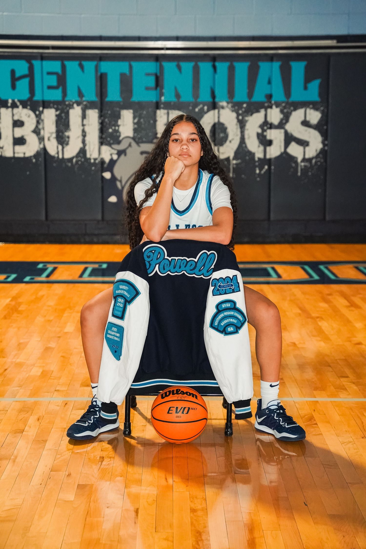Basketball player poses on gym floor with Centennial Bulldogs backdrop wearing team uniform and holding a basketball.
