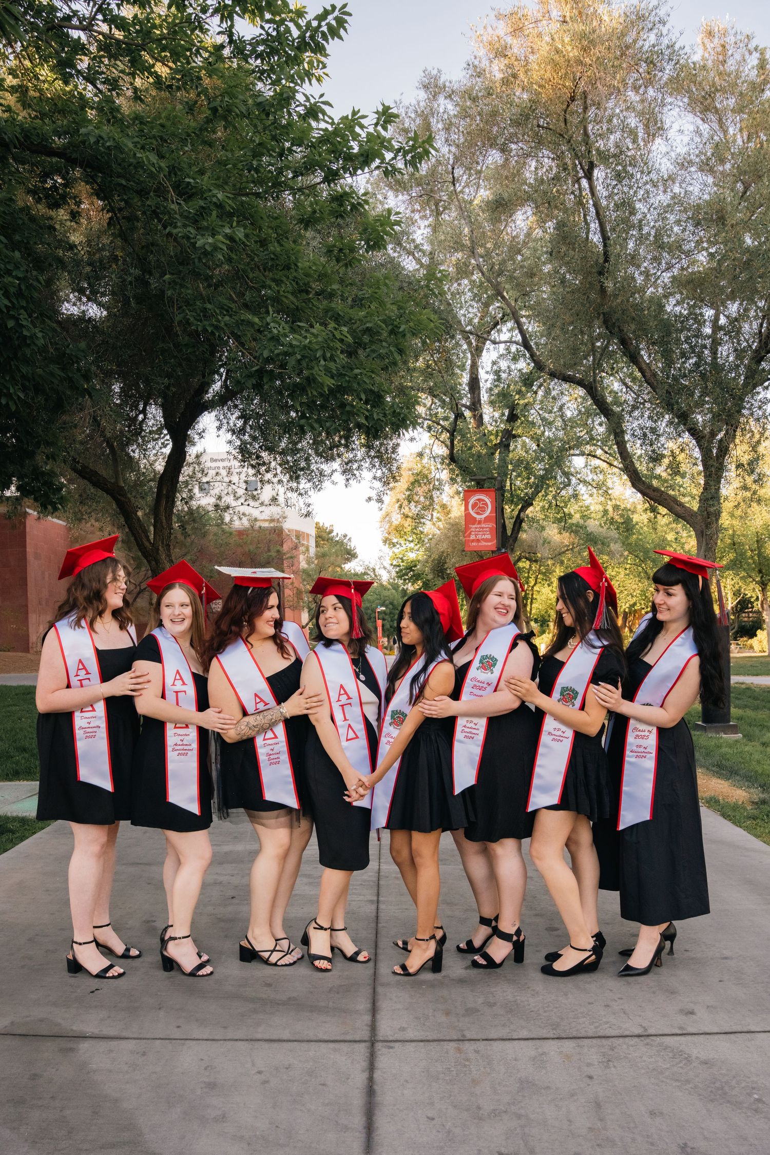 Graduates in black dresses with pink sashes pose casually in groups on campus grounds.
