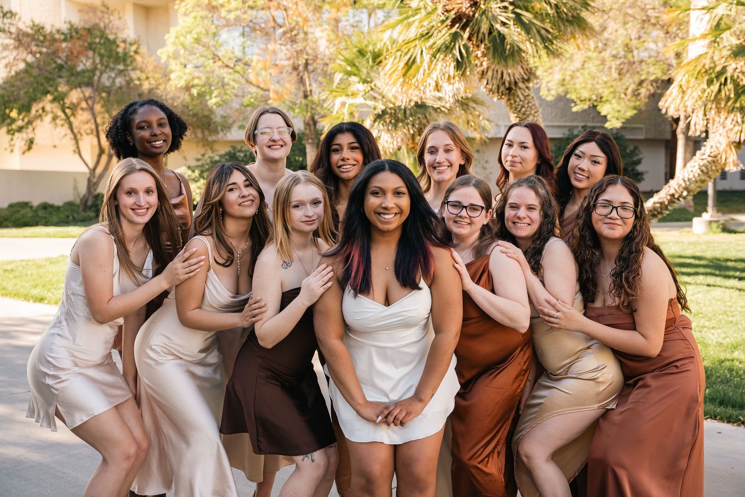 Large group of friends in neutral dresses pose together outdoors in a garden setting.