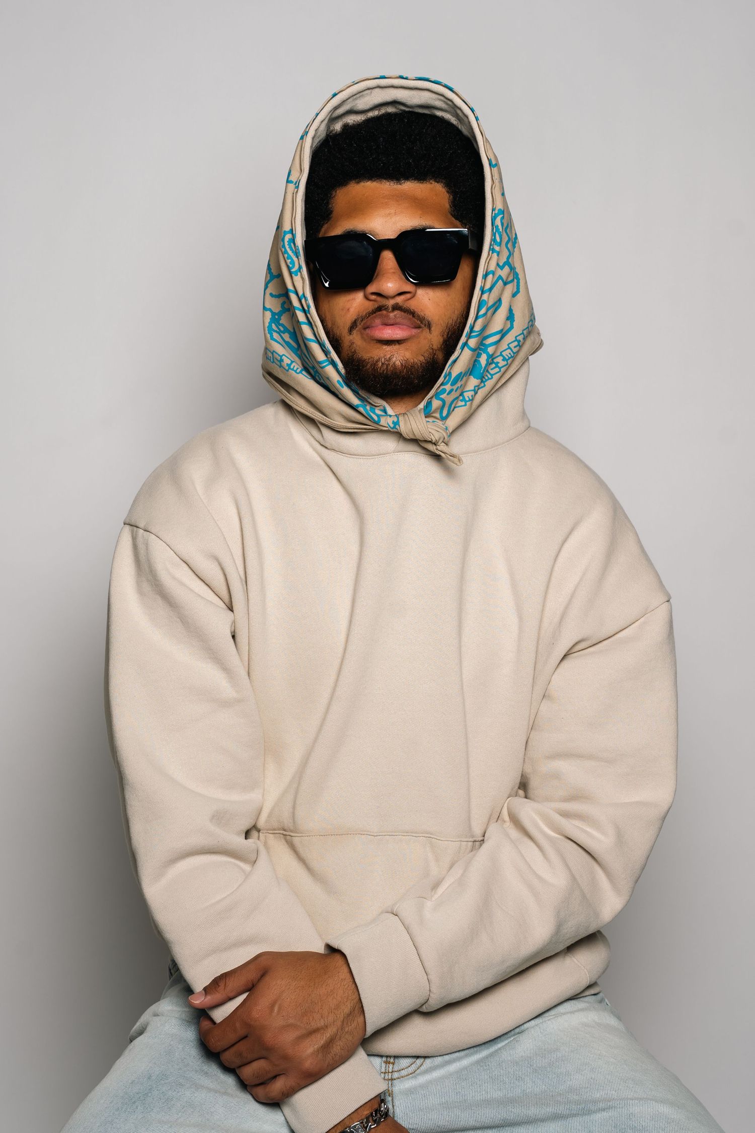 Monaco Media. Fashion portrait of person in cream hoodie with patterned headscarf and sunglasses against gray background.