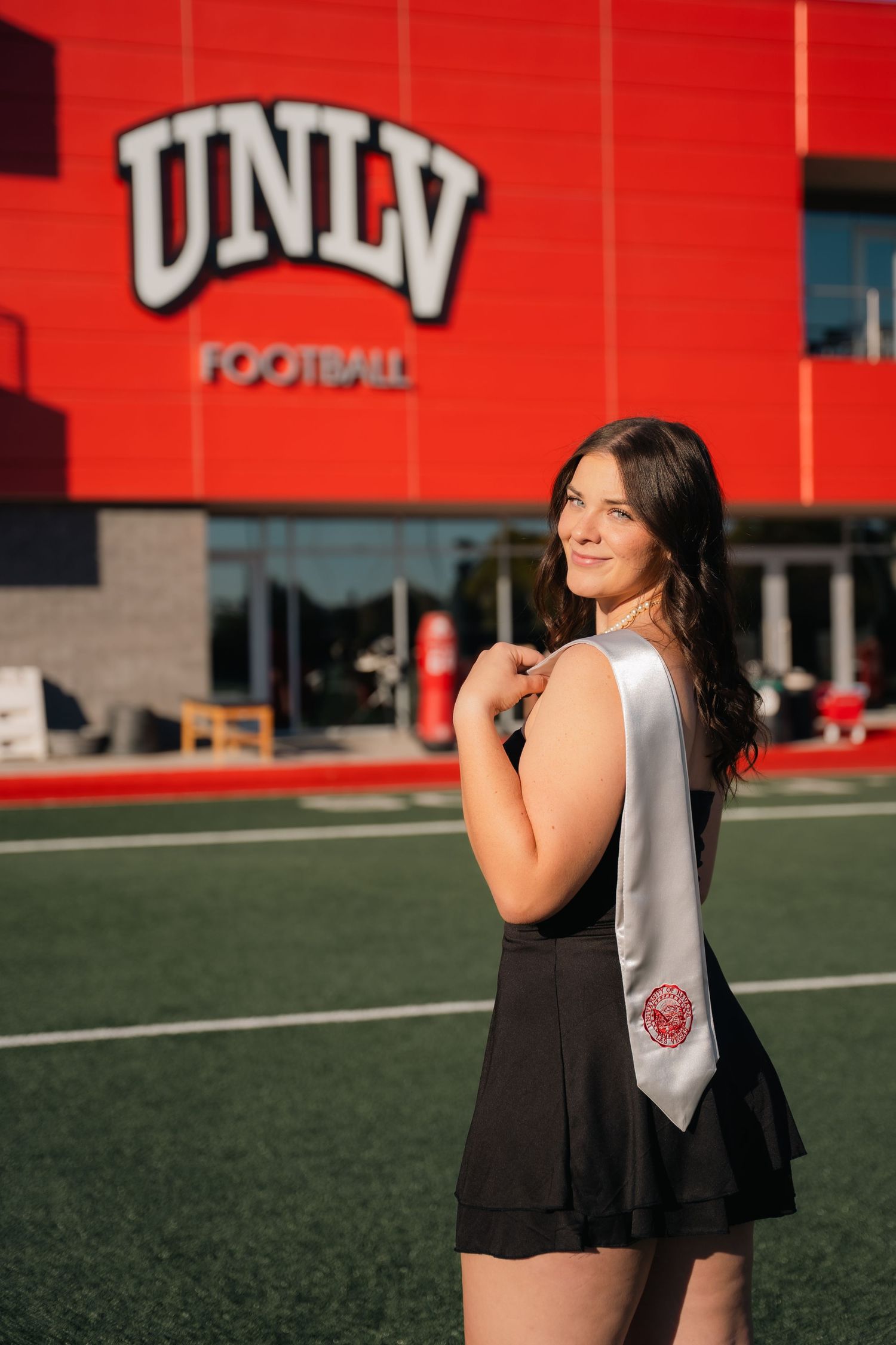 Graduate wearing graduation stole poses in black dress at UNLV football facility.
