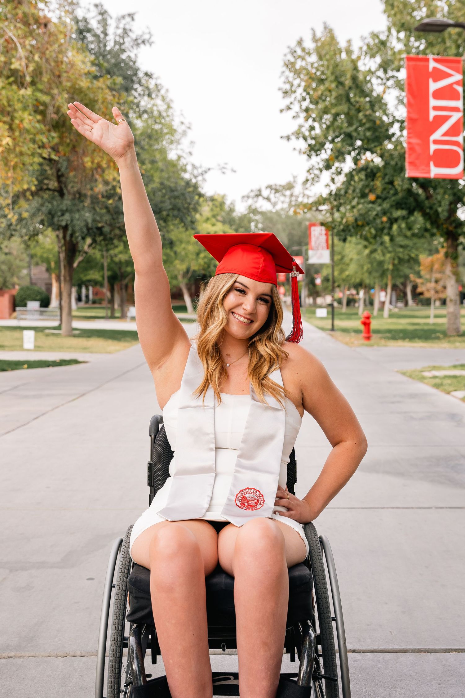 Graduate in wheelchair celebrating with raised arm in white dress and red cap on university campus.
