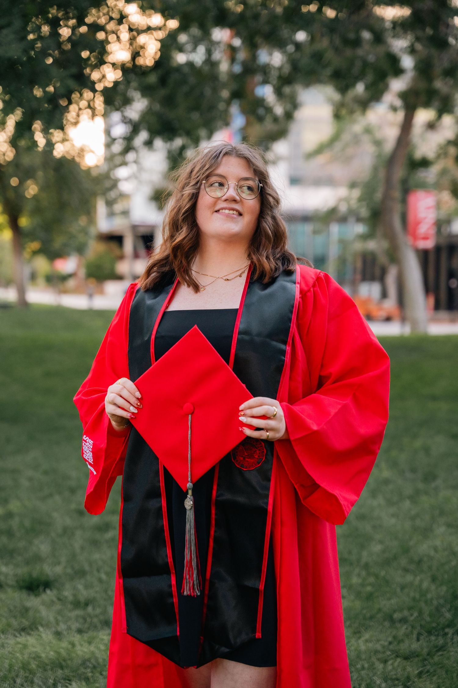 Graduate in red cap and gown poses joyfully on grassy campus grounds.