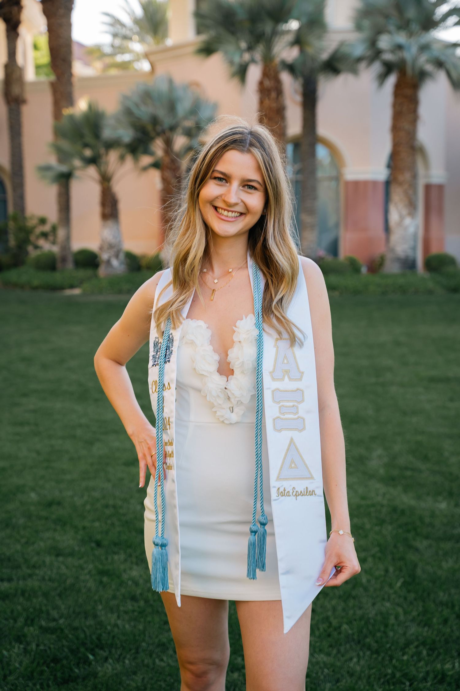Graduate wearing white dress and light blue graduation stole poses outdoors by palm trees and pink building.