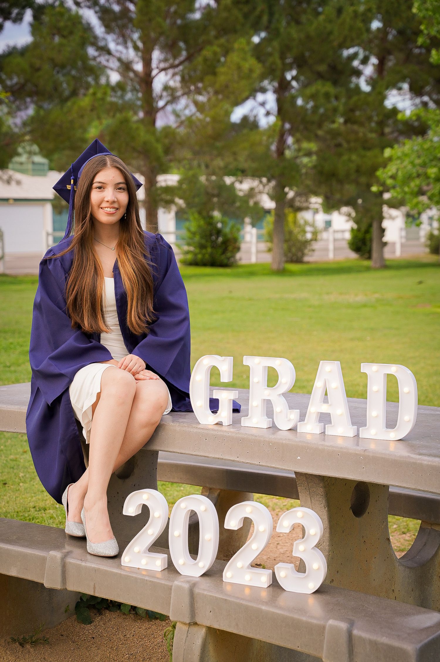 Graduate in purple cap and gown sits on bench with GRAD 2023 letters for graduation photos on campus.