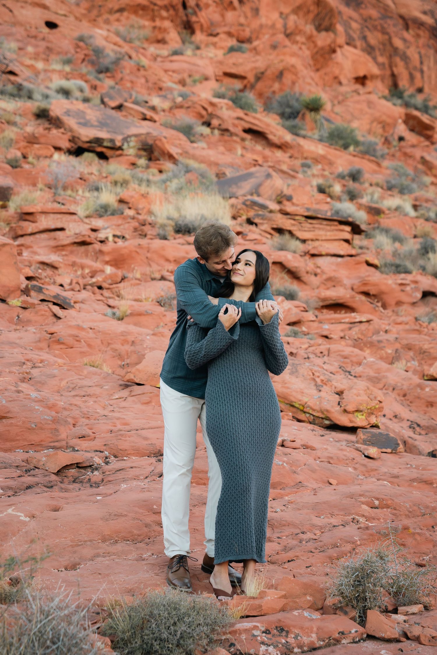 A couple sharing an intimate embrace on red desert rocks with desert brush in the background.