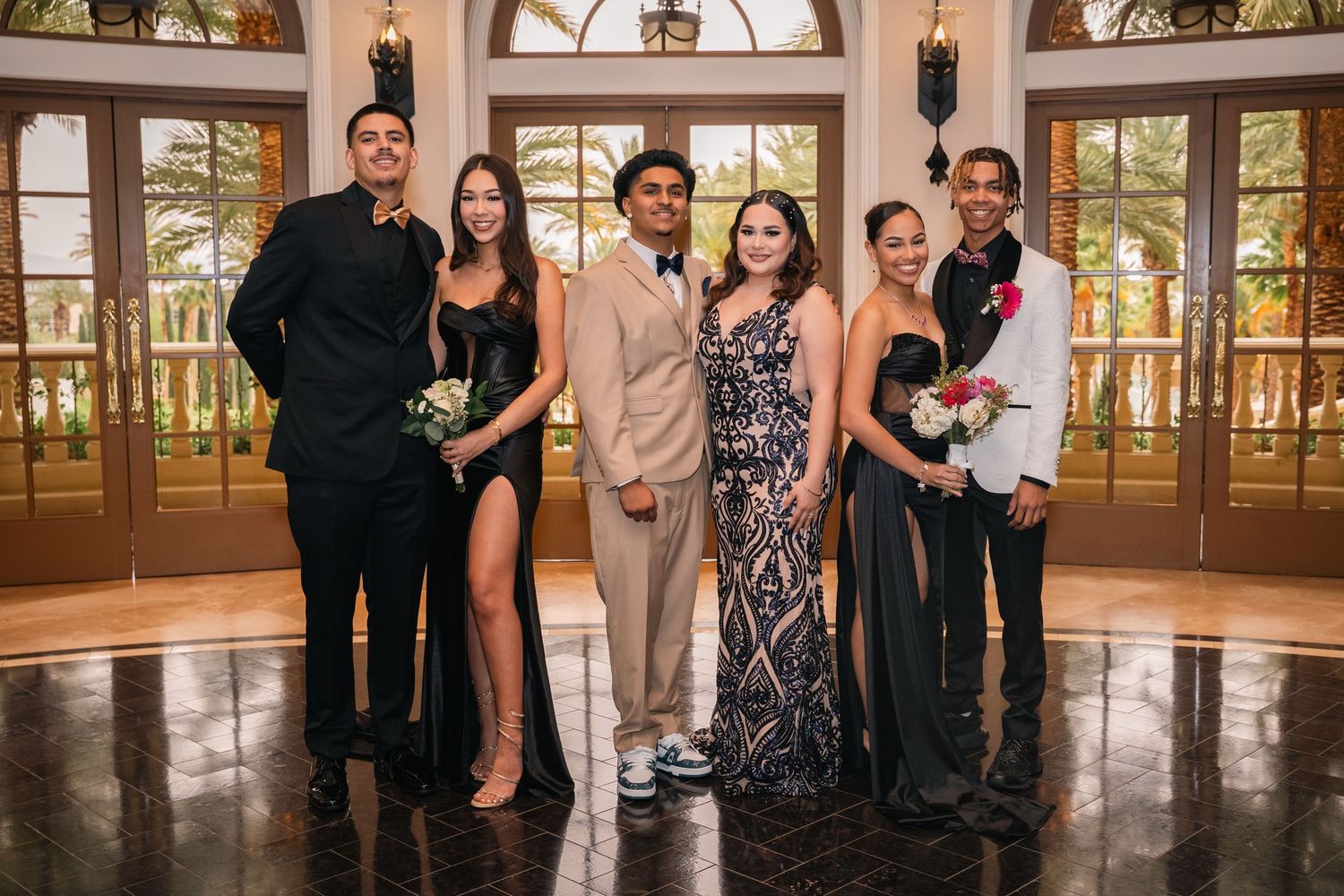 Three formal couples pose together at an elegant venue with arched windows and ornate decor.